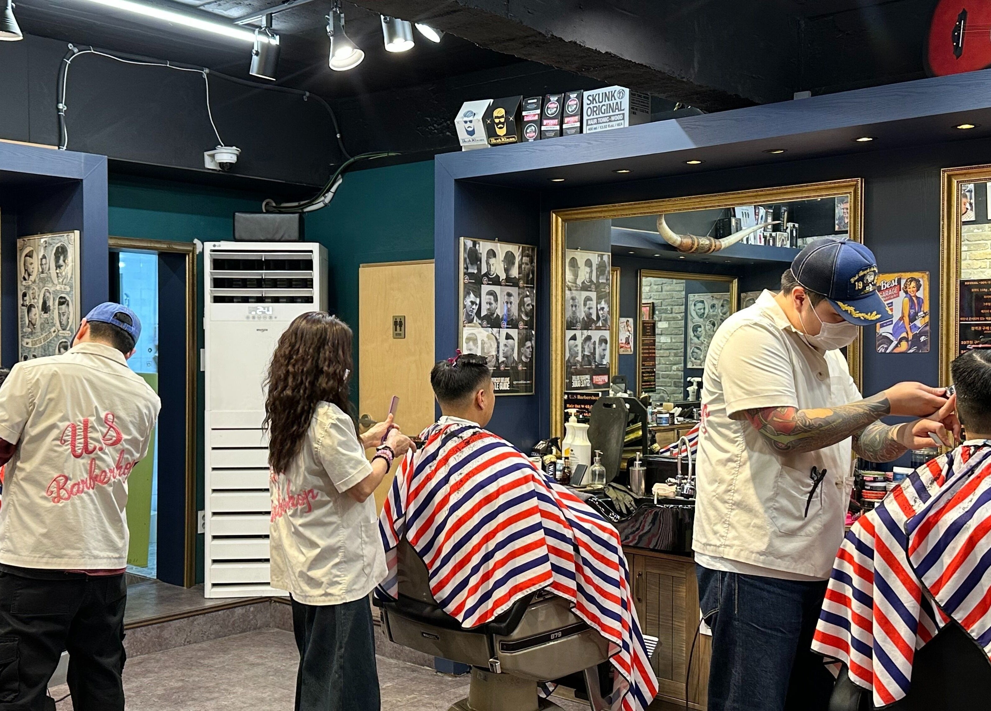 Stylists at US Barbershop, 서울특별시, KR, trim hair under modern lighting with red-white-blue capes.