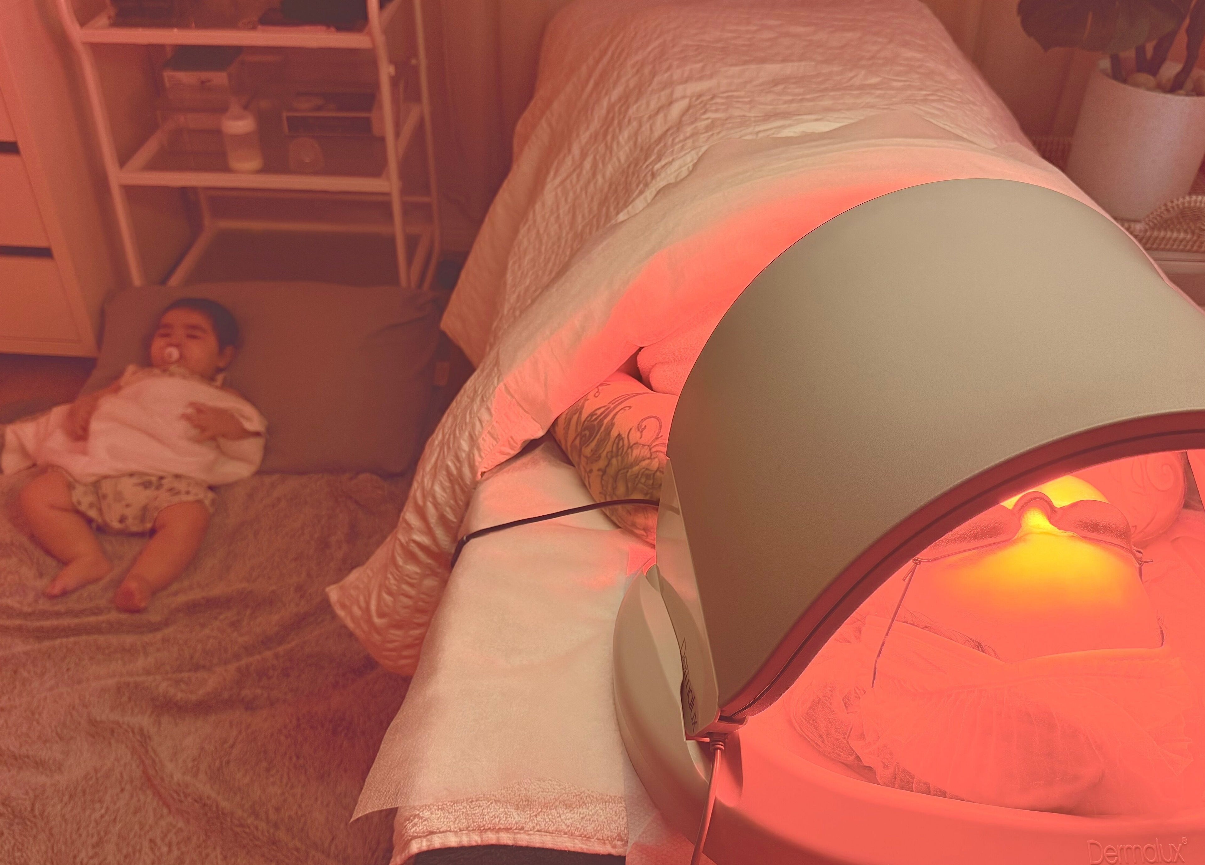 Cozy wellness setup at M.N.M Beauty Bar, Roselands, NSW. A person under LED treatment beside a resting baby.