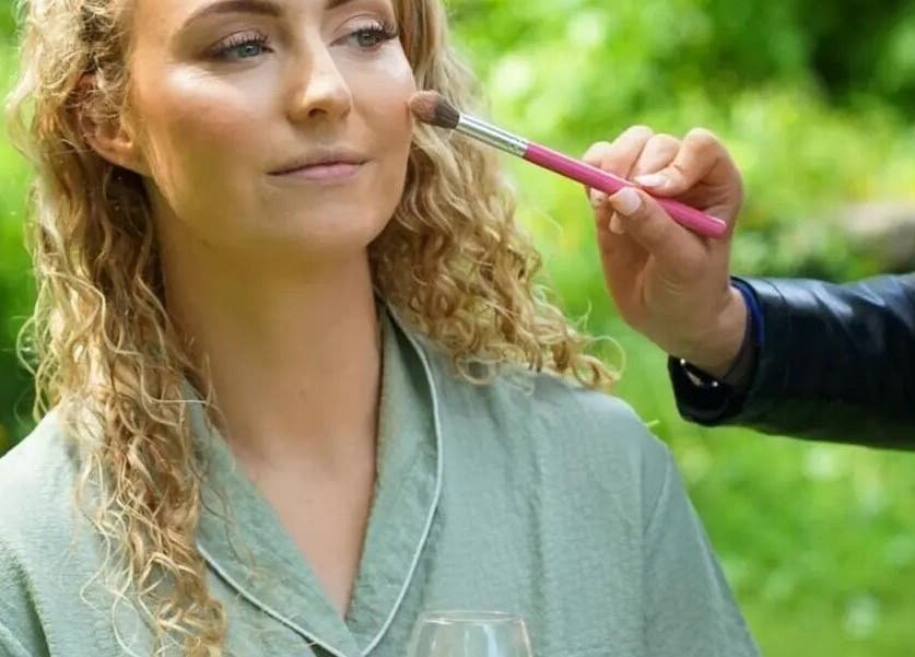 Makeup artist applies blush at Kerie Beauty, Saint Asaph, Wales, GB, enhancing a woman's complexion outdoors.