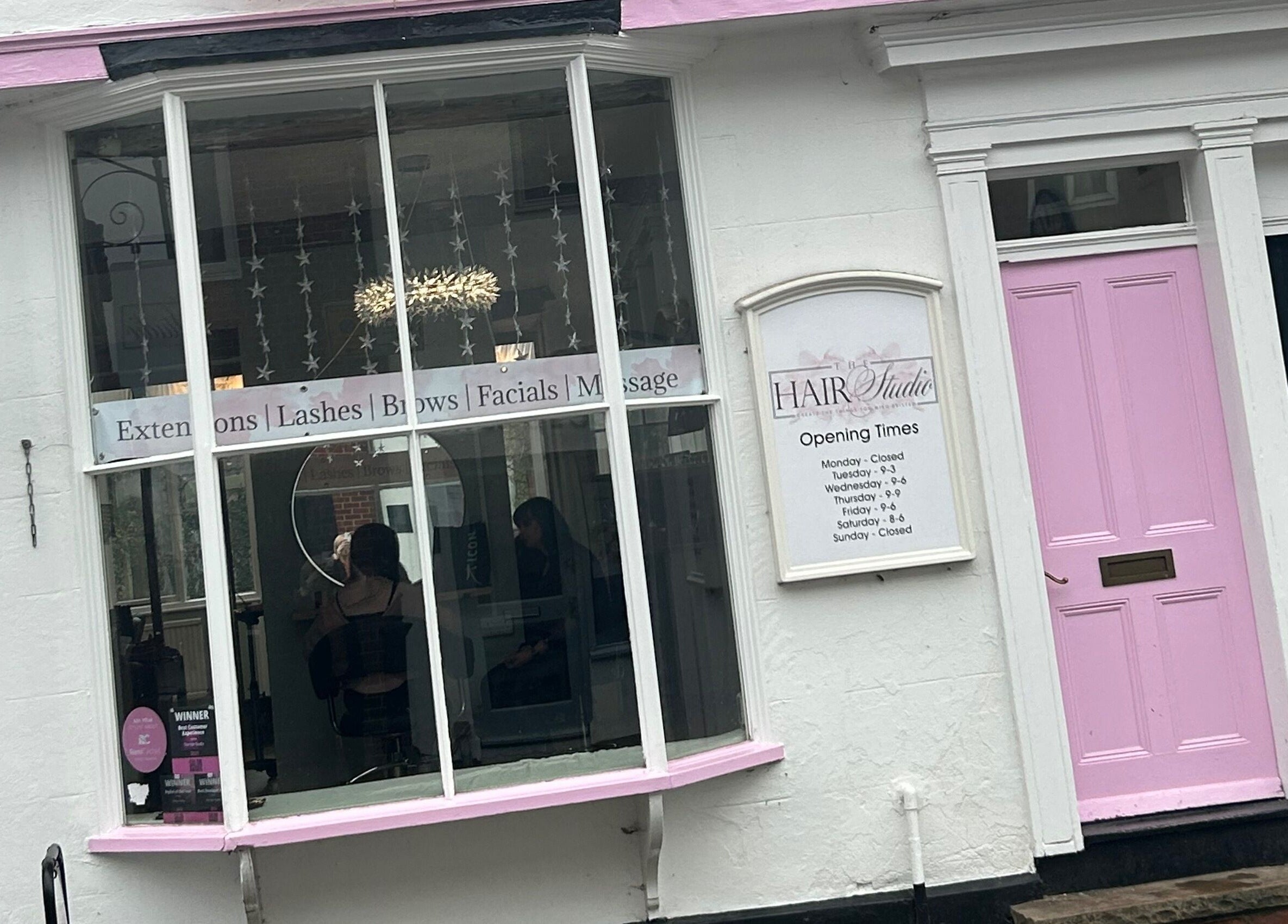 Front view of The Hair Studio in Sudbury, England, GB with a pink door and services displayed in the window.