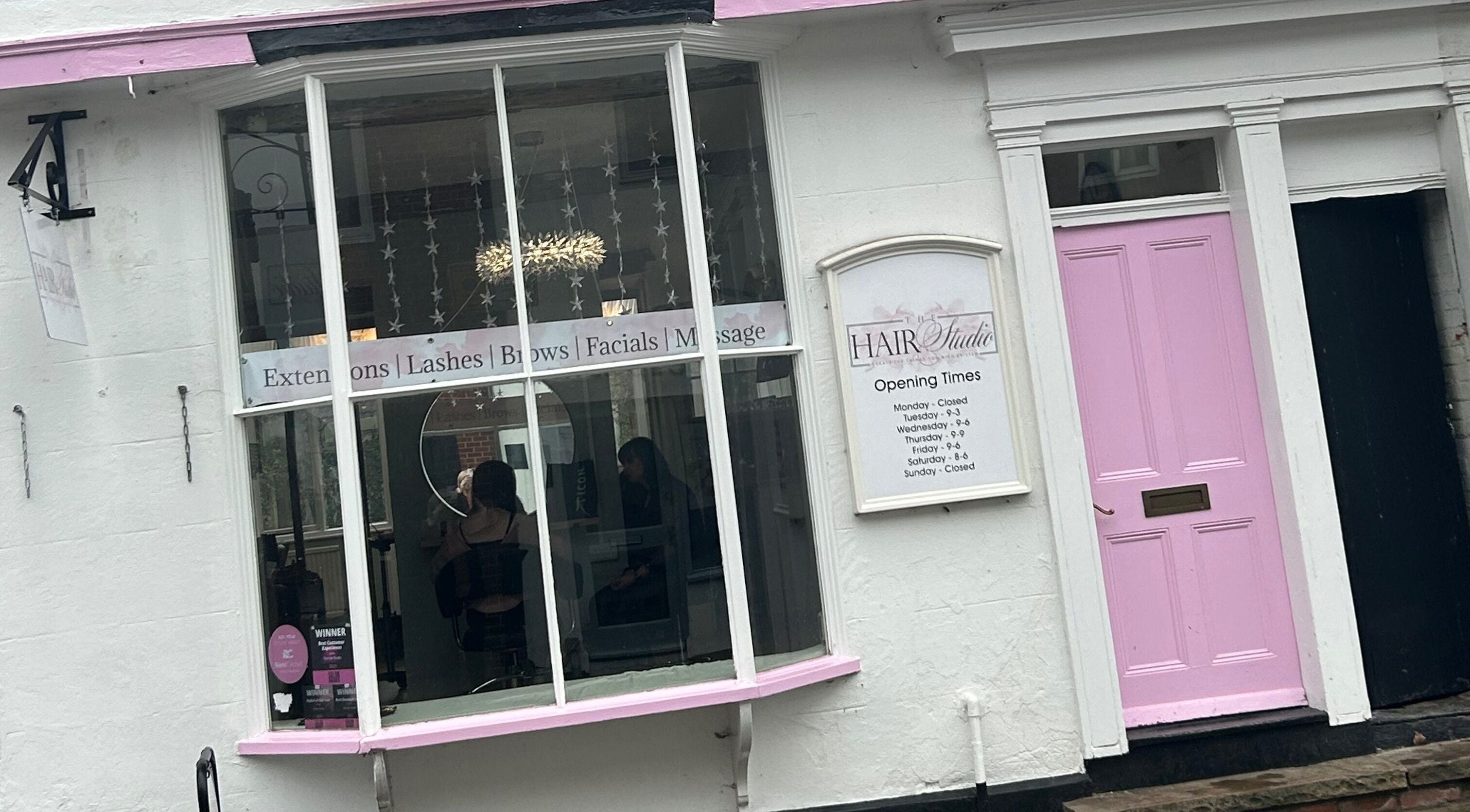 Front view of The Hair Studio in Sudbury, England, GB with a pink door and services displayed in the window.
