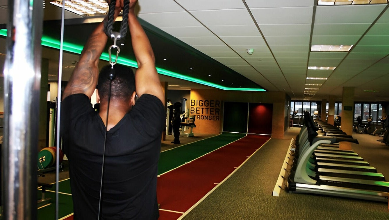 A gym-goer using equipment at Direct Gainz in Birmingham, England, GB, with treadmills and fitness area visible.