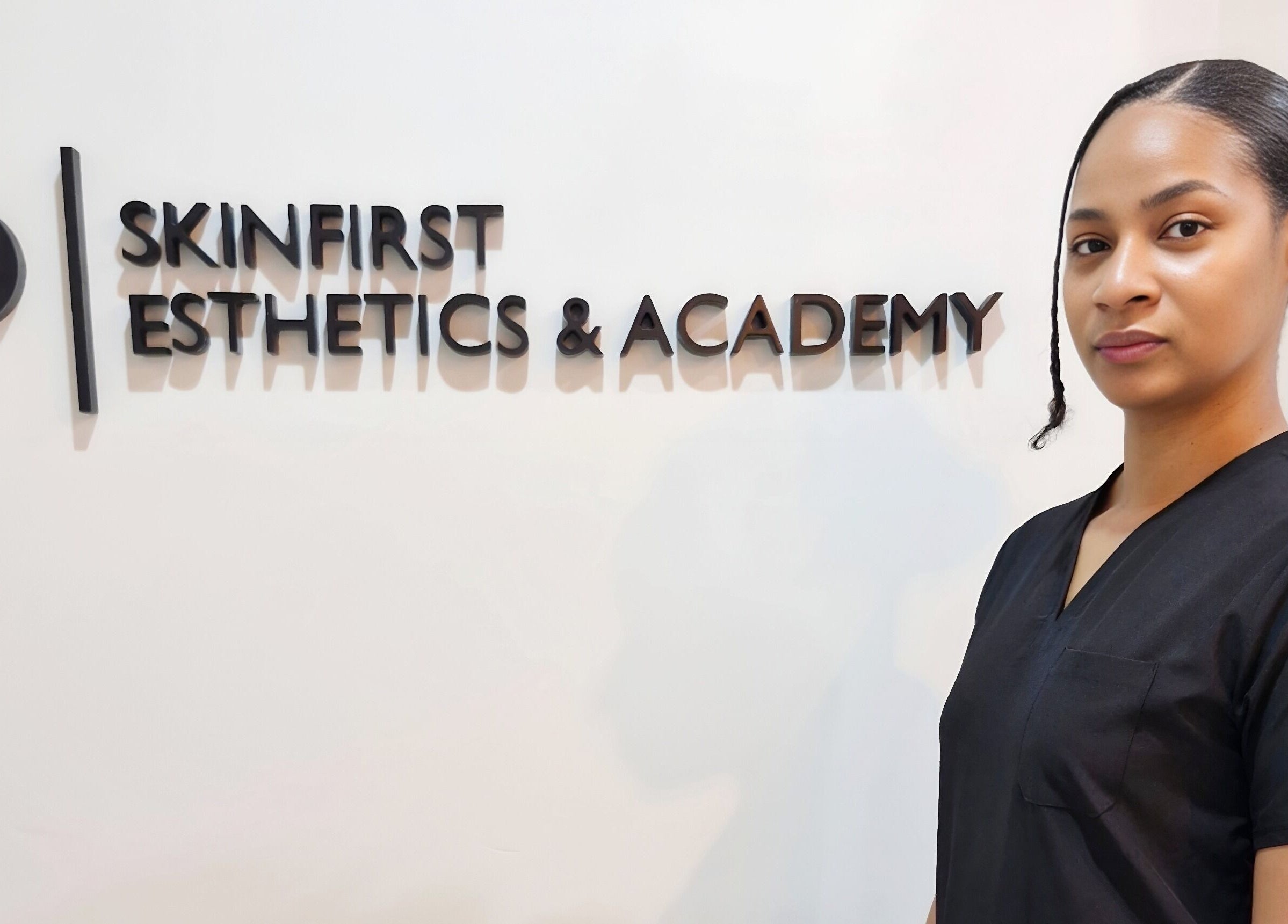 Staff member at SkinFirst Esthetics & Academy in La Tourney, Vieux Fort, LC beside logo wall.