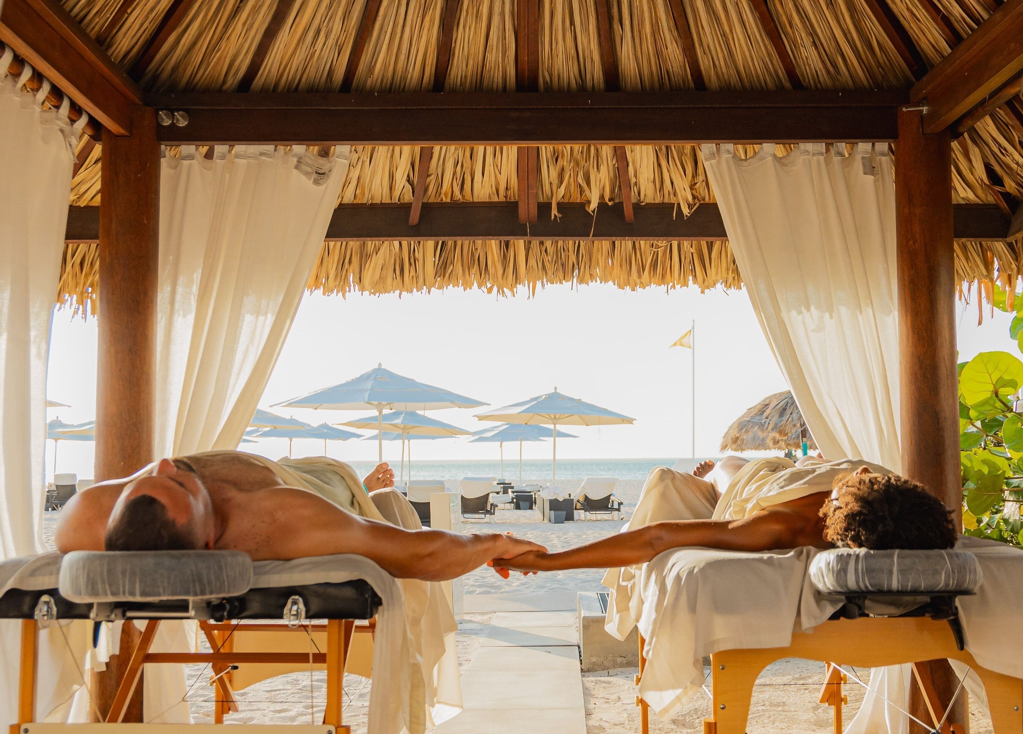 Couple enjoys beachfront massages at Purun Spa, Oranjestad, AW under a tiki hut with ocean view.