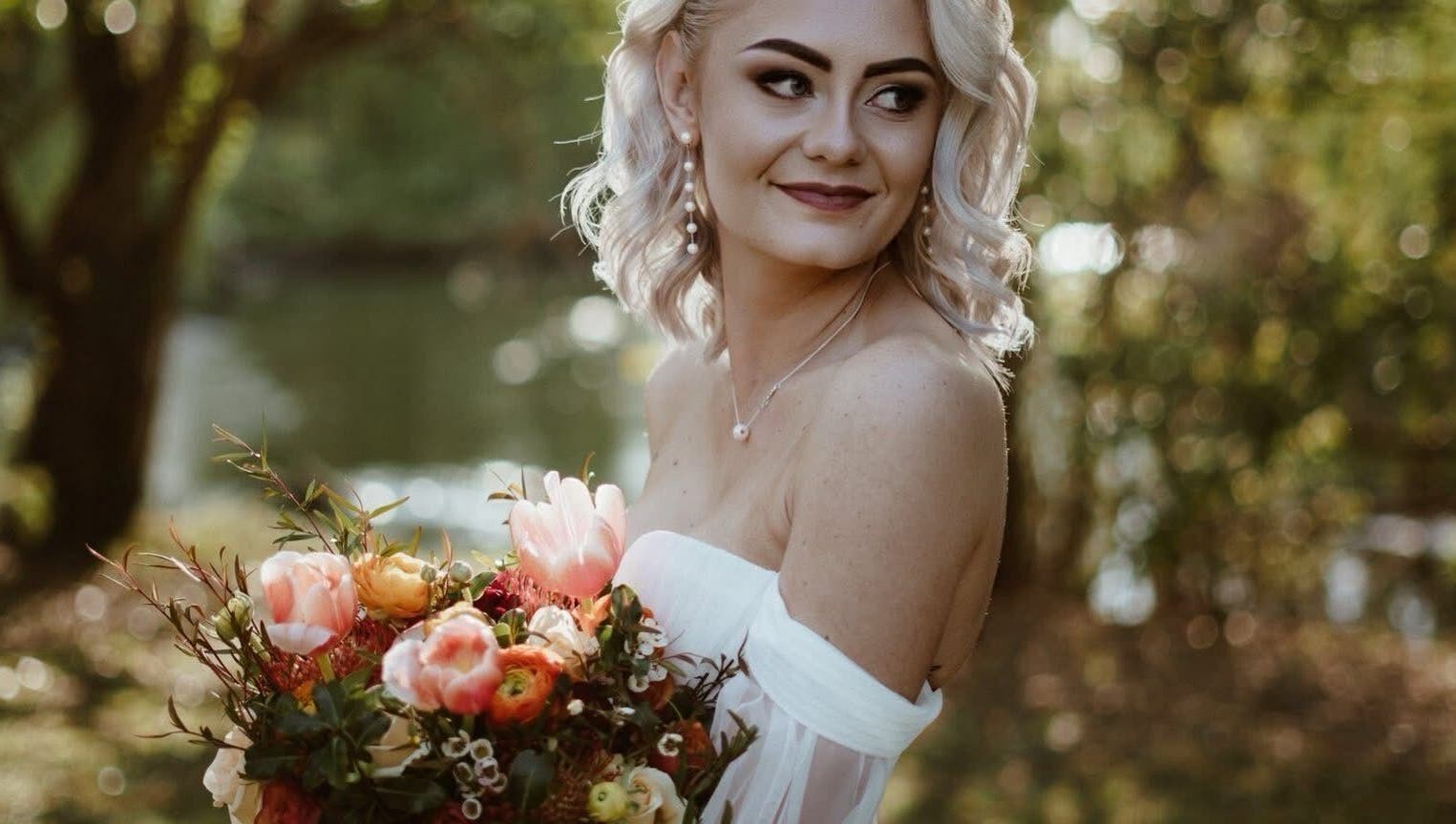 A smiling bride with a bouquet at Skoonveldt, Klerksdorp, North West, ZA, surrounded by lush greenery.