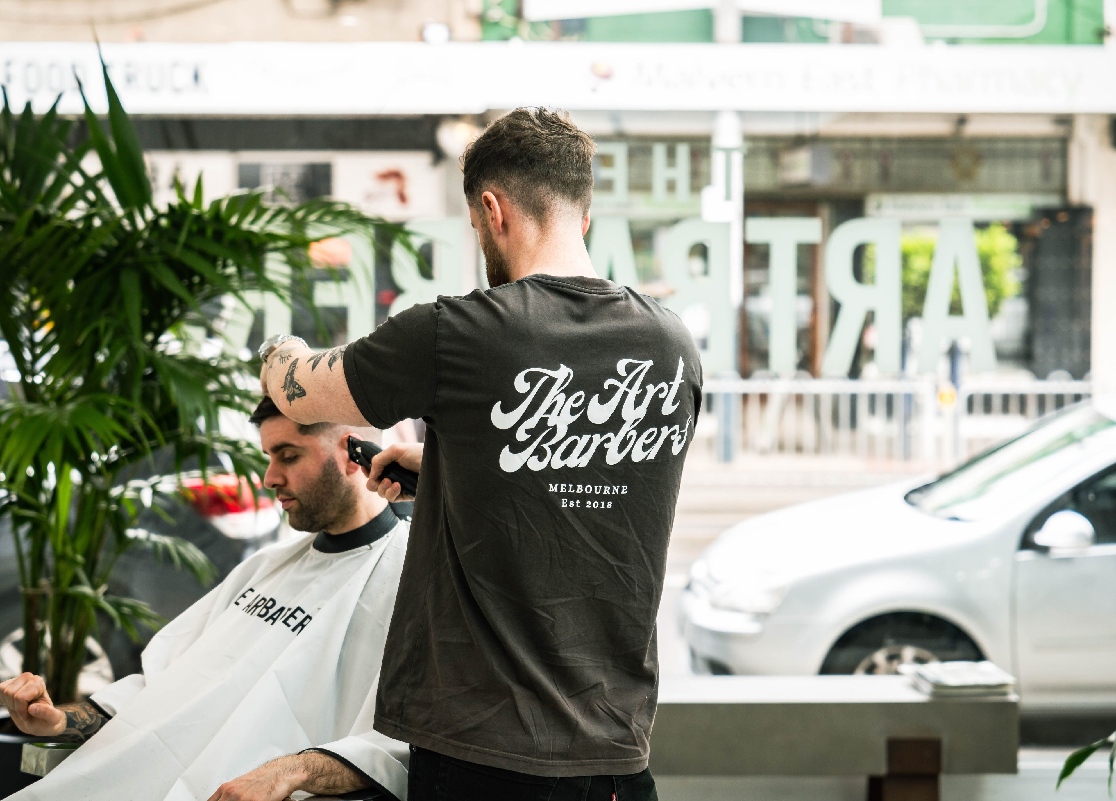 Barber trimming hair at The Art Barbers Malvern in Melbourne, Victoria, AU with plants and street view.