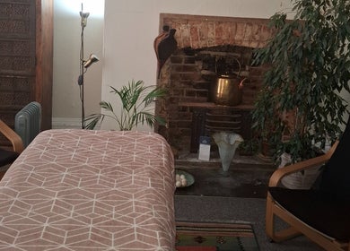 Relaxation room at Shass Therapy Massage, Cheltenham, England featuring a massage table, armchair, and brick fireplace.