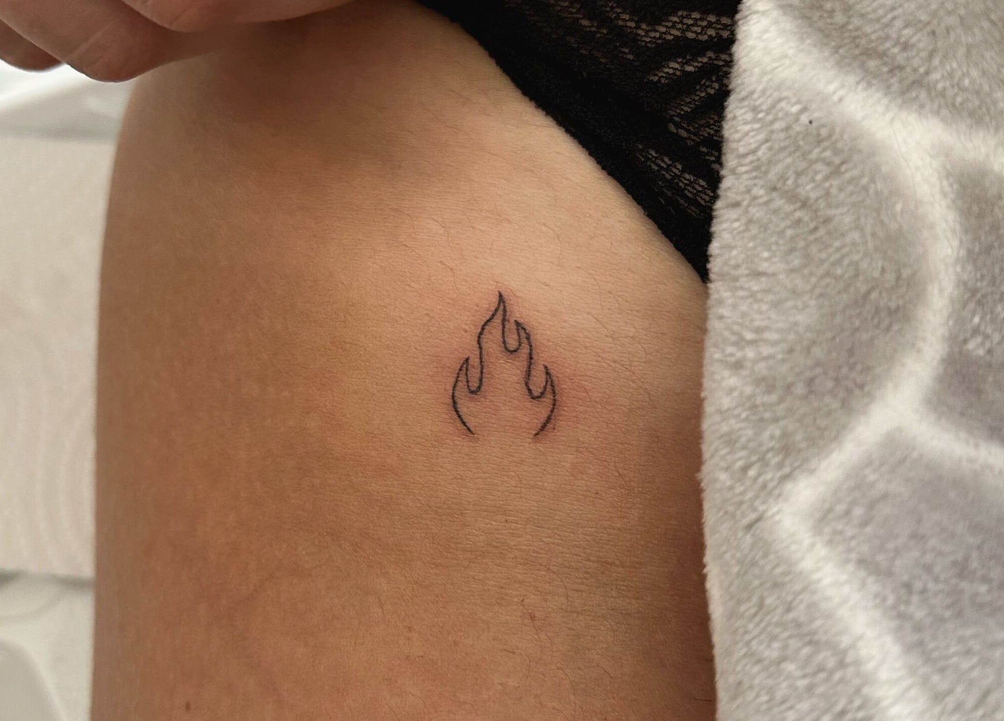 Small flame tattoo on skin at Indybeautyuk, Stilton, England, GB. Fine line detail enhances its artistic design.