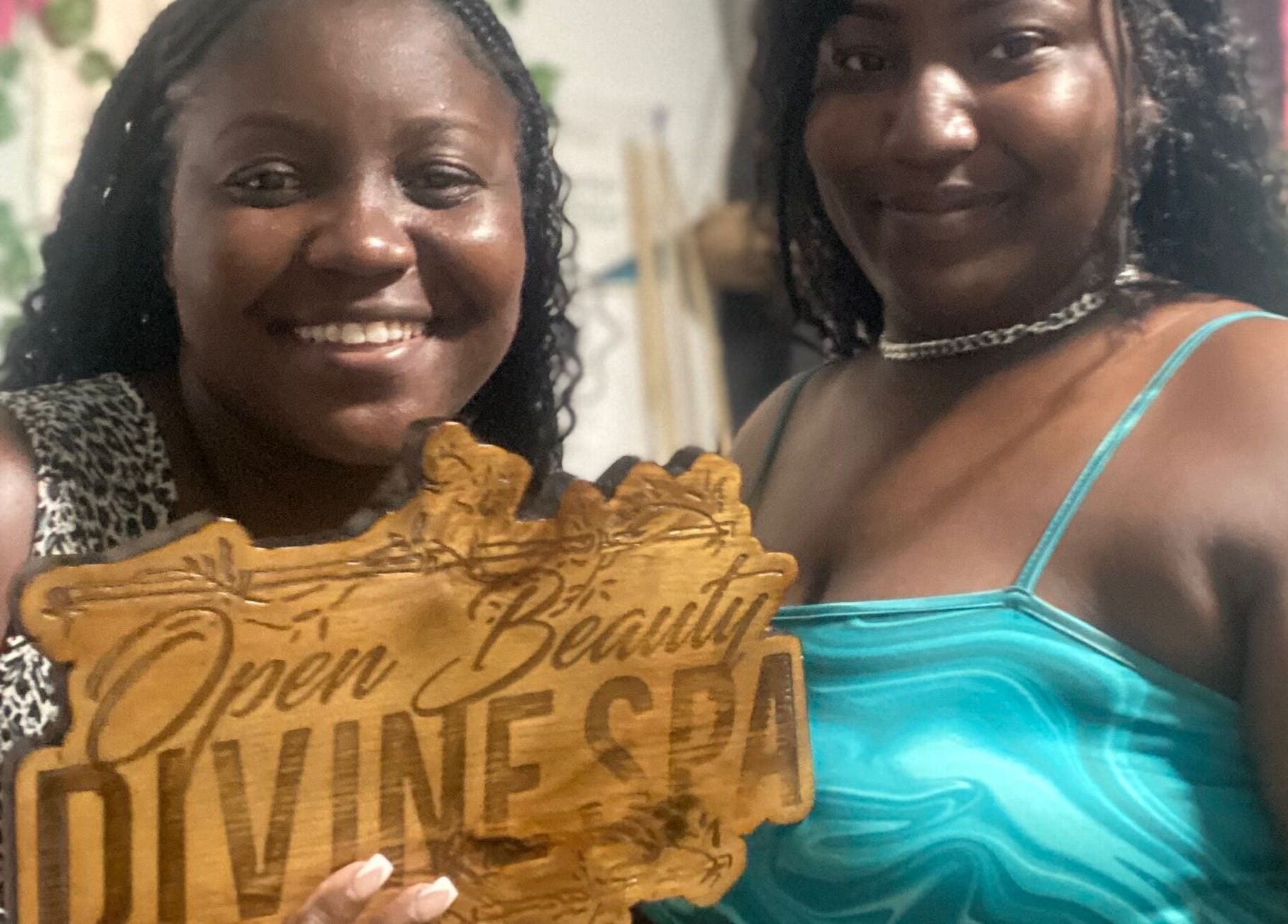 Two smiling women hold Open Beauty Divine Spa sign at Kingston, St. Andrew Parish, JM.