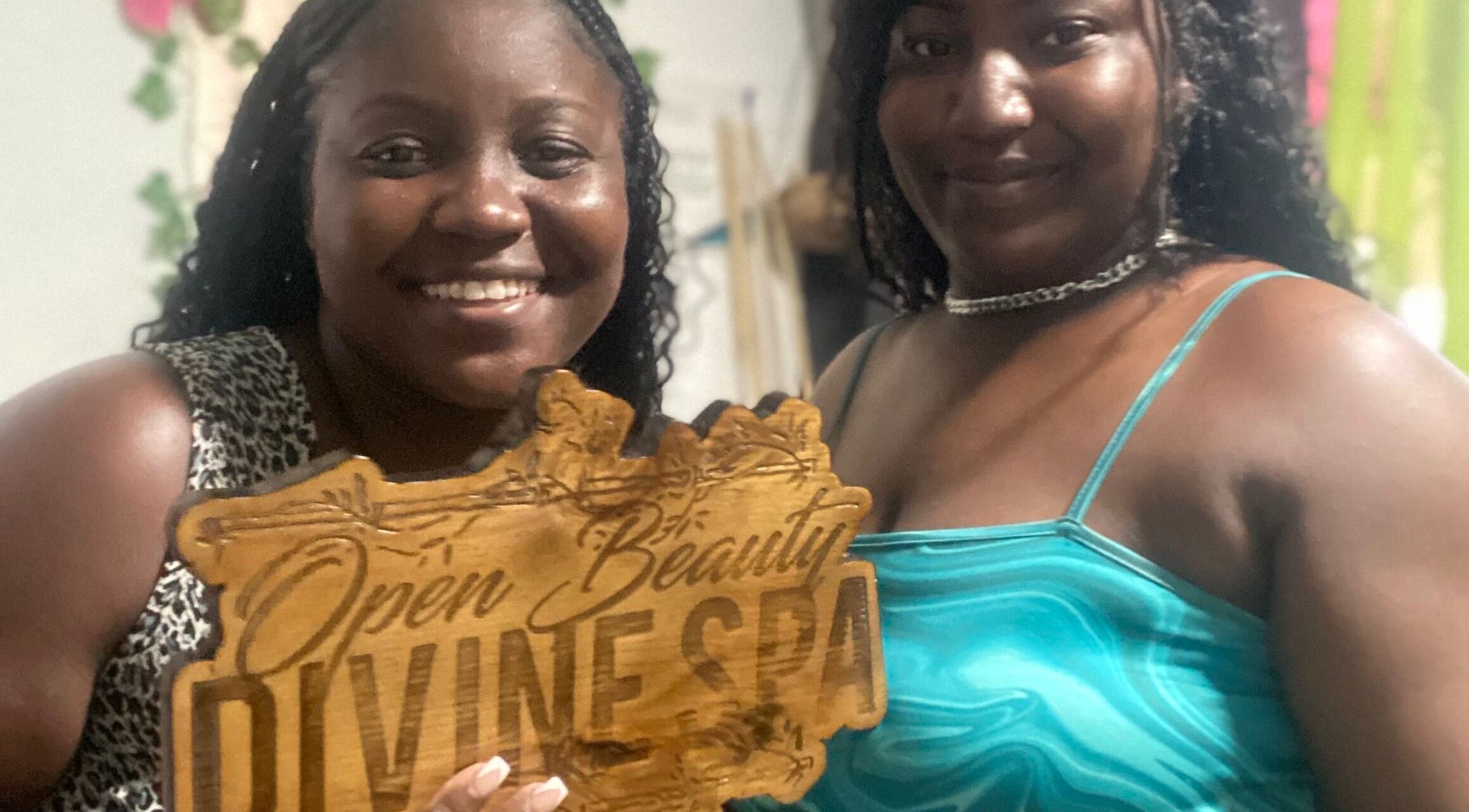 Two smiling women hold Open Beauty Divine Spa sign at Kingston, St. Andrew Parish, JM.