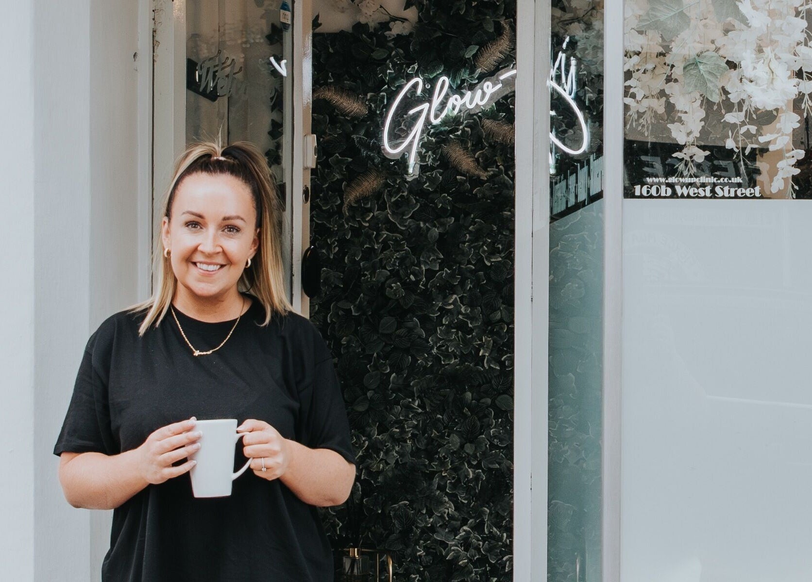 Smiling person at The Glow Up Clinic entrance, Fareham, England, GB with plants and neon sign.