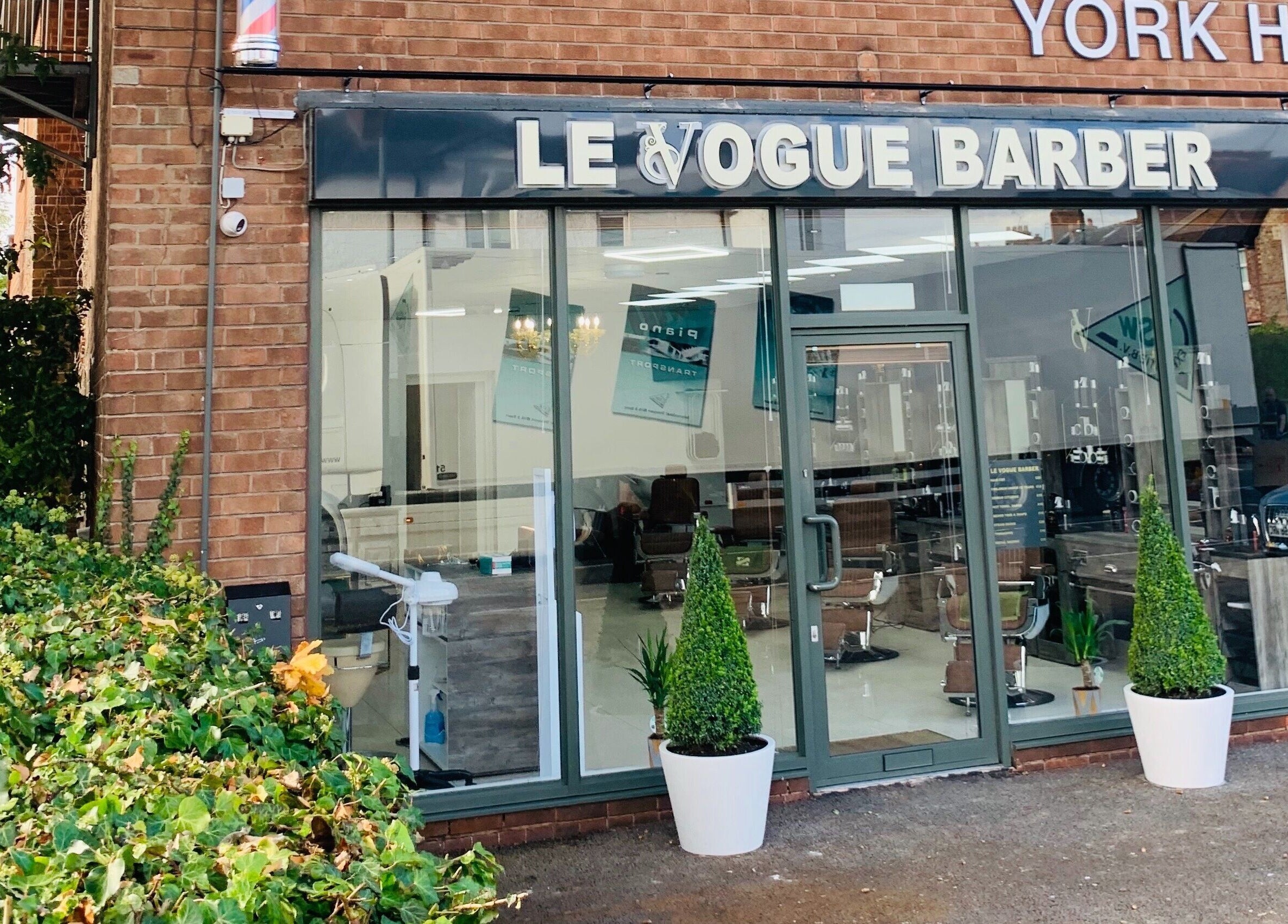 Exterior view of Le Vogue Barber in Bowdon, England, GB featuring modern glass facade and greenery.