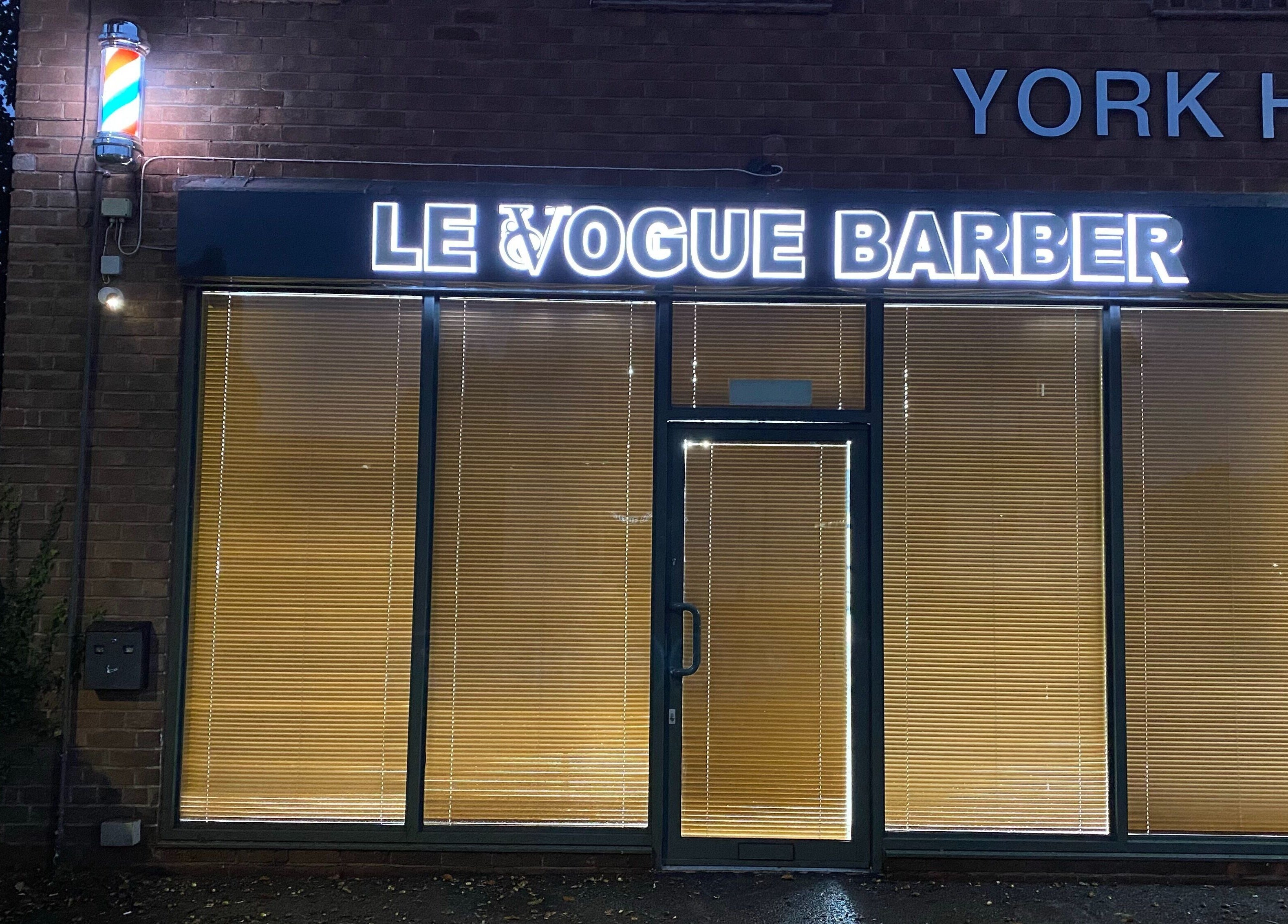 Le Vogue Barber facade at Bowdon, England, GB, displaying illuminated signage and classic barber pole.
