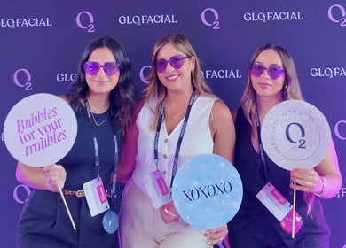 Three women at the Glo2 event at Bliss Spa & Wellness, San Juan, posing with fun signs and purple sunglasses.