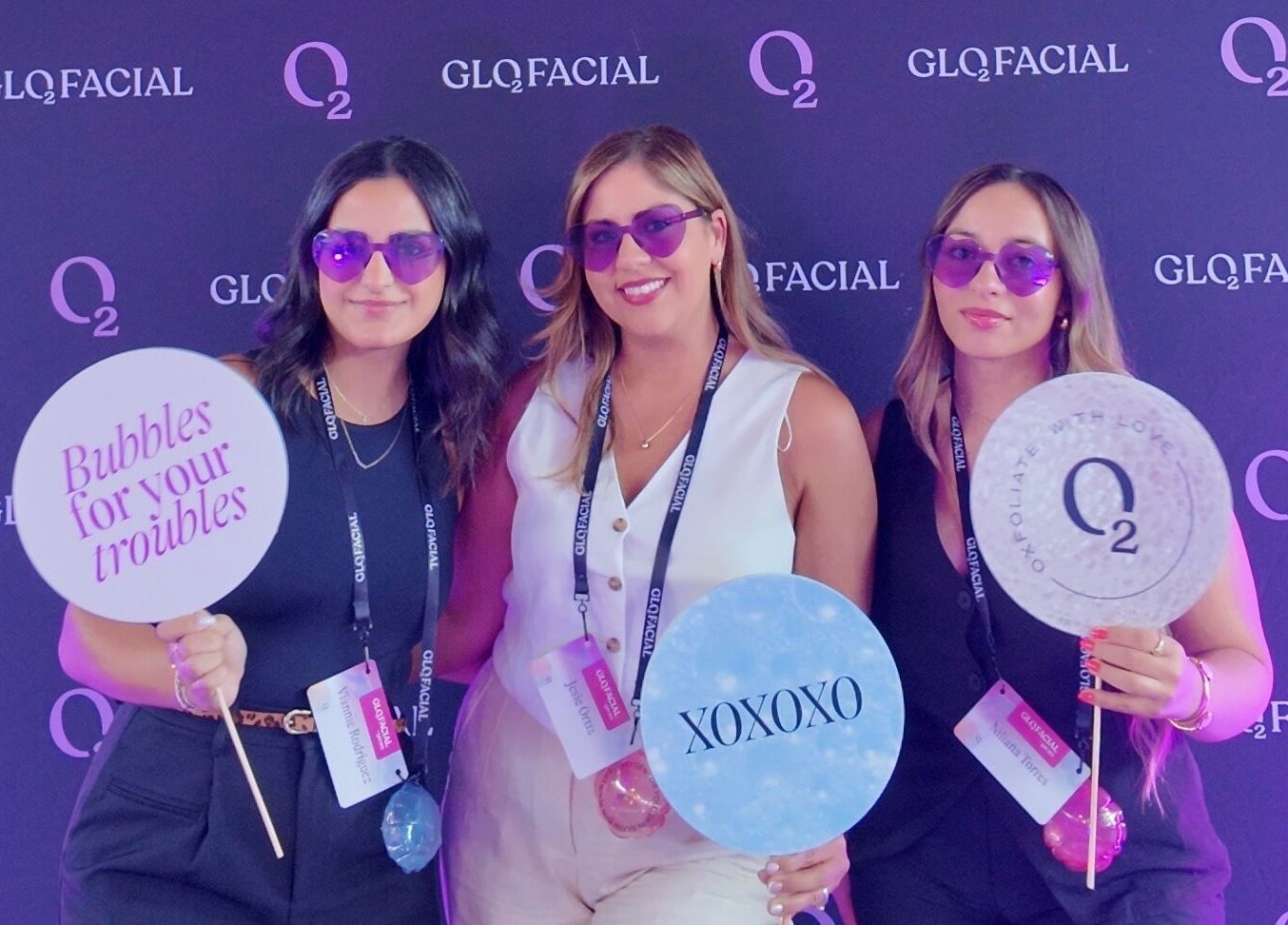 Three women at the Glo2 event at Bliss Spa & Wellness, San Juan, posing with fun signs and purple sunglasses.