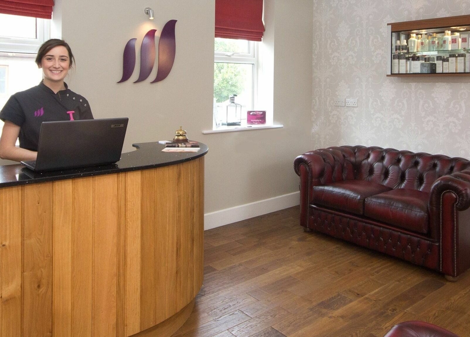 Reception area at New Image in Bangor, Wales, GB featuring a smiling staff member, wooden desk, and leather sofa.