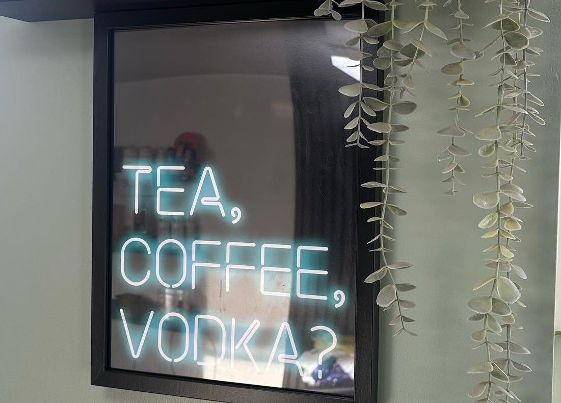A neon sign at Envious Salon, Crawley, England, GB stating 'Tea, Coffee, Vodka?' with greenery beside it.