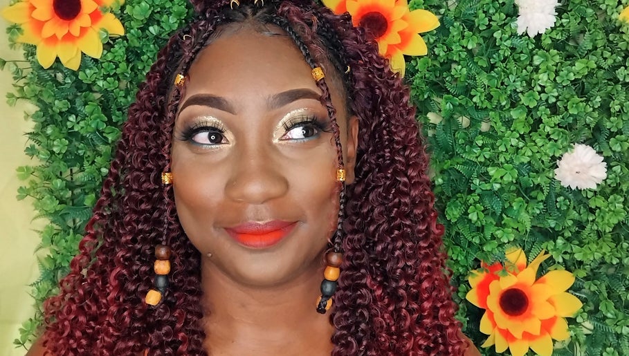 A woman with vibrant makeup and braided curls smiles with floral backdrop at Niva Creations, Saint Michael, BB.