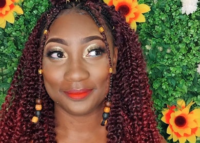 A woman with vibrant makeup and braided curls smiles with floral backdrop at Niva Creations, Saint Michael, BB.