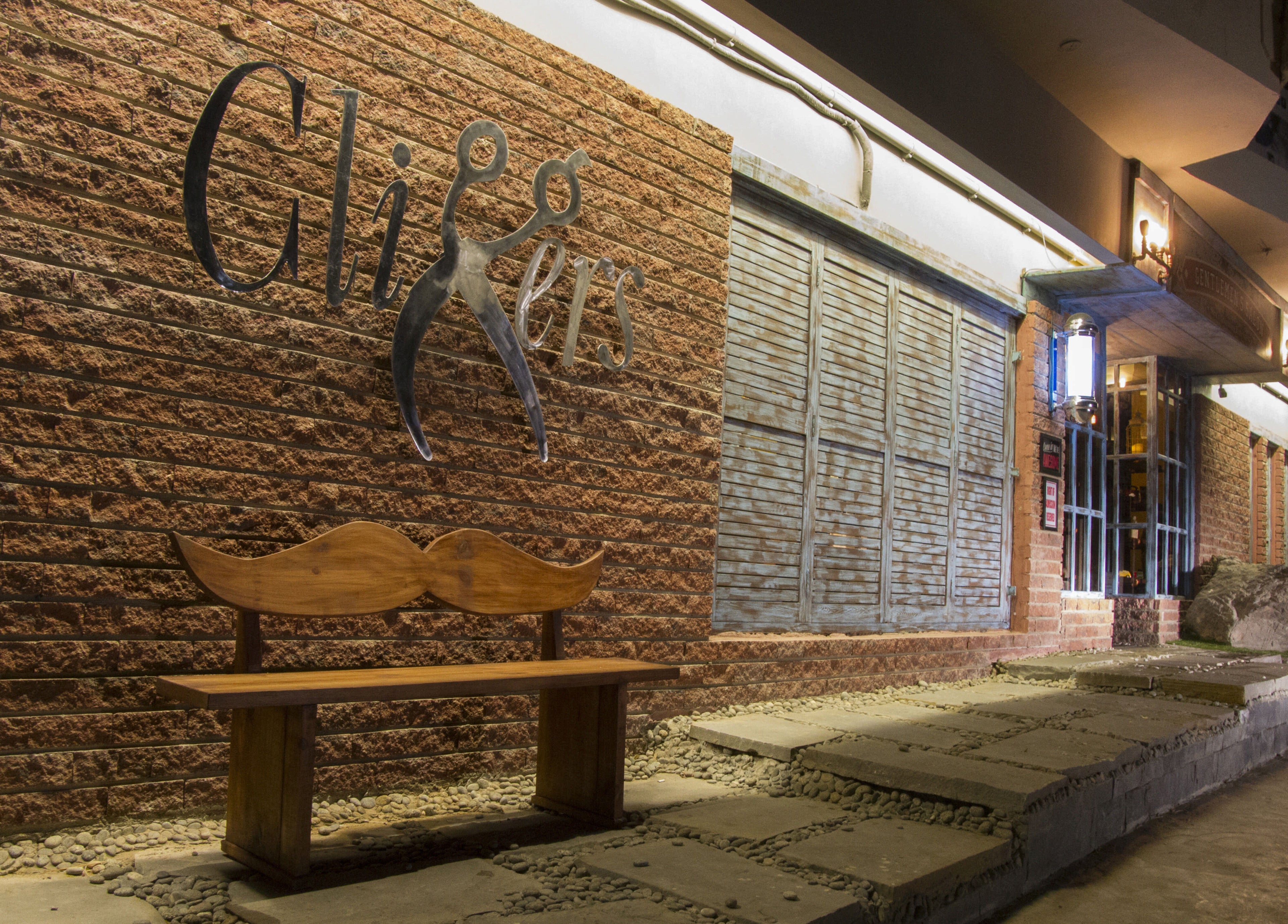Front view of Clippers Salon Zamzama Branch, Karachi, Sindh, PK, with brick wall and mustache bench.