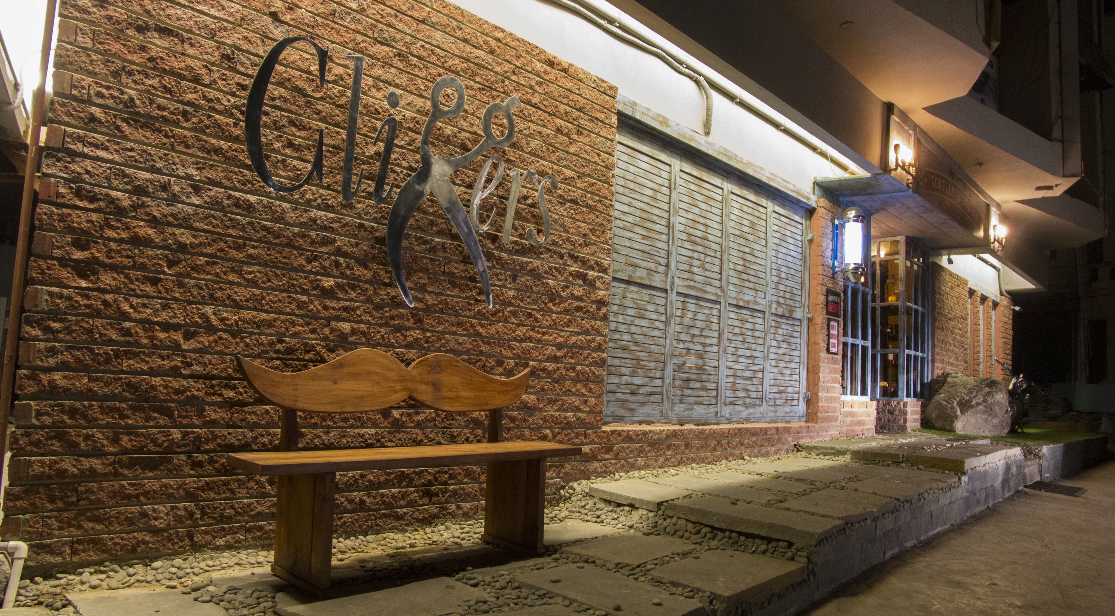 Front view of Clippers Salon Zamzama Branch, Karachi, Sindh, PK, with brick wall and mustache bench.