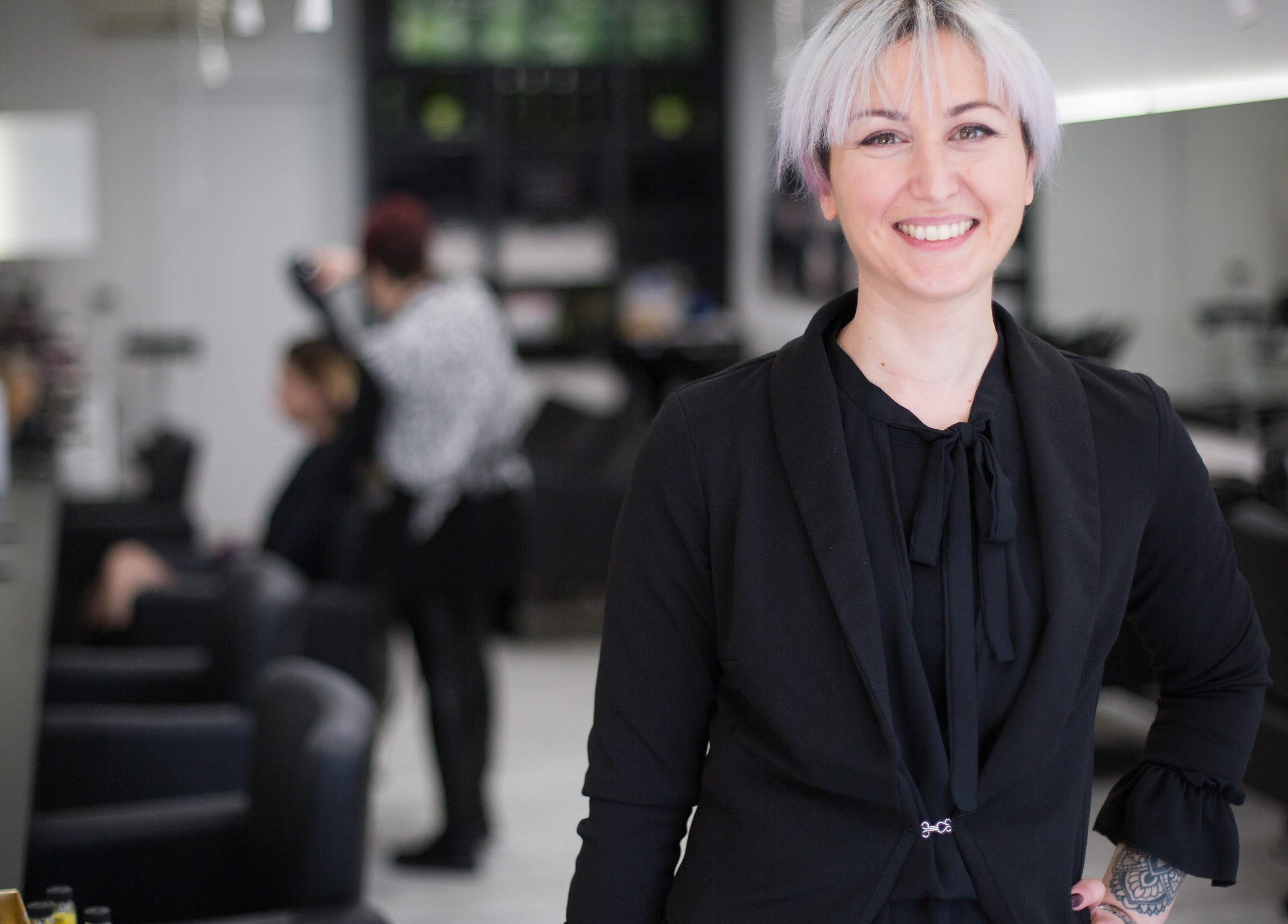 Stilista sorridente presso Elegance Parrucchieri, Pescara, Italia, IT, con clienti sullo sfondo.