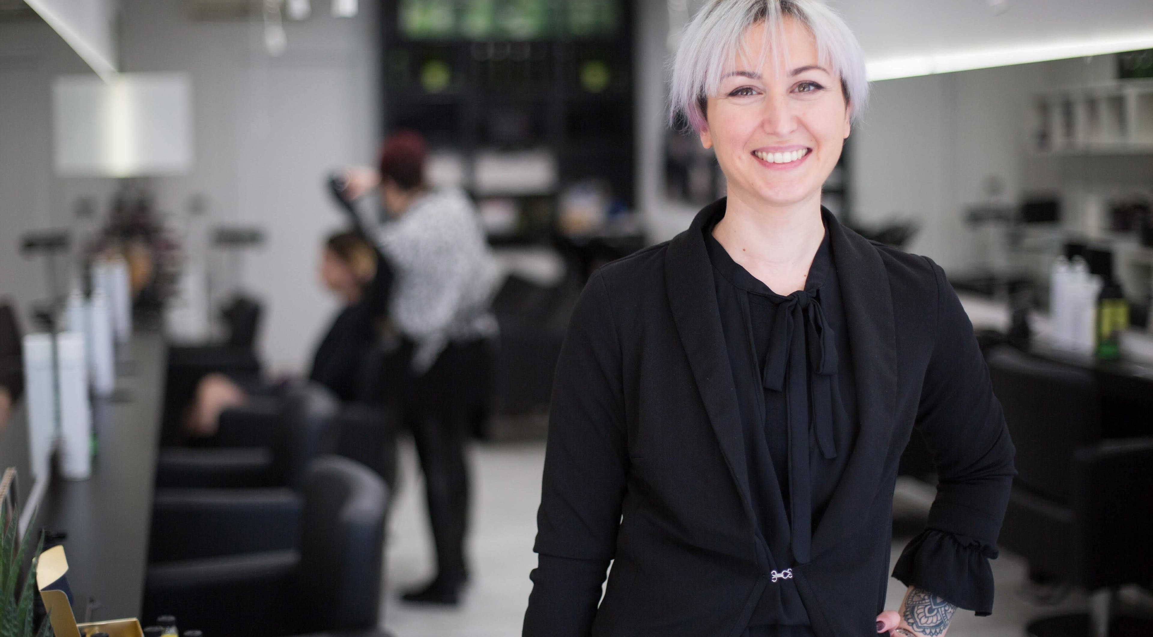 Stilista sorridente presso Elegance Parrucchieri, Pescara, Italia, IT, con clienti sullo sfondo.