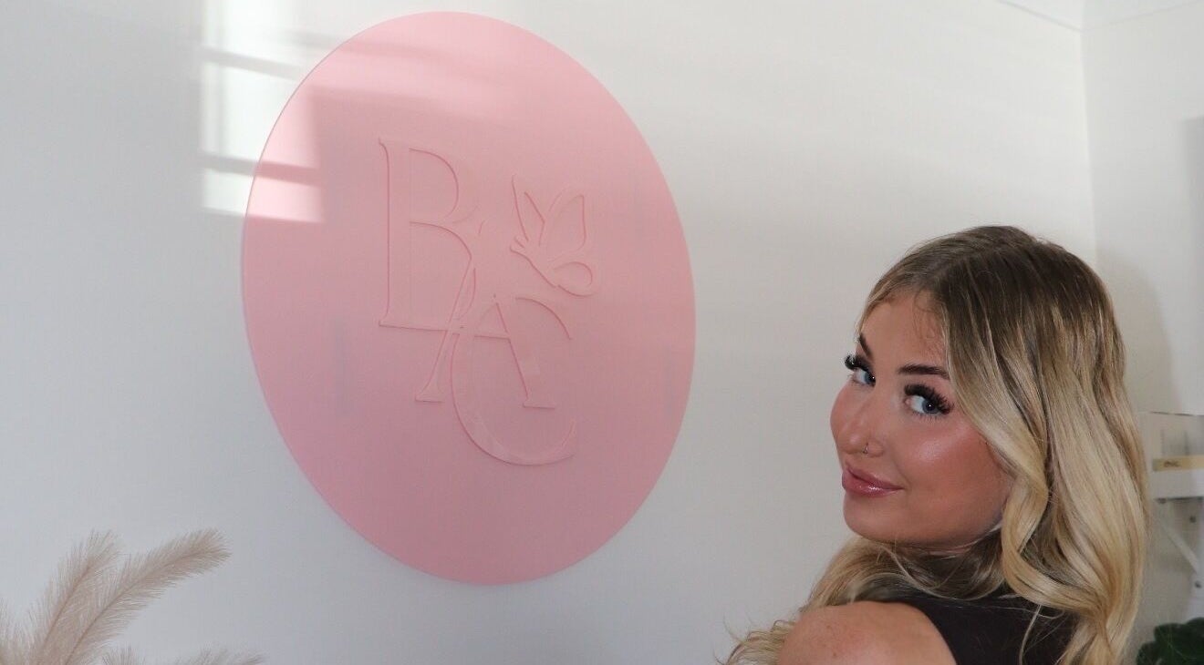 Smiling client at Beauty at Caitlyn’s - Glen Alpine Beauty Hub, Glen Alpine, New South Wales, AU beside pink logo.