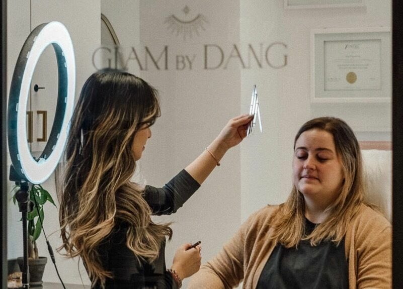 Expert completes eyebrow styling at Glam by Dang, New York, NY with a focused client under a ring light.