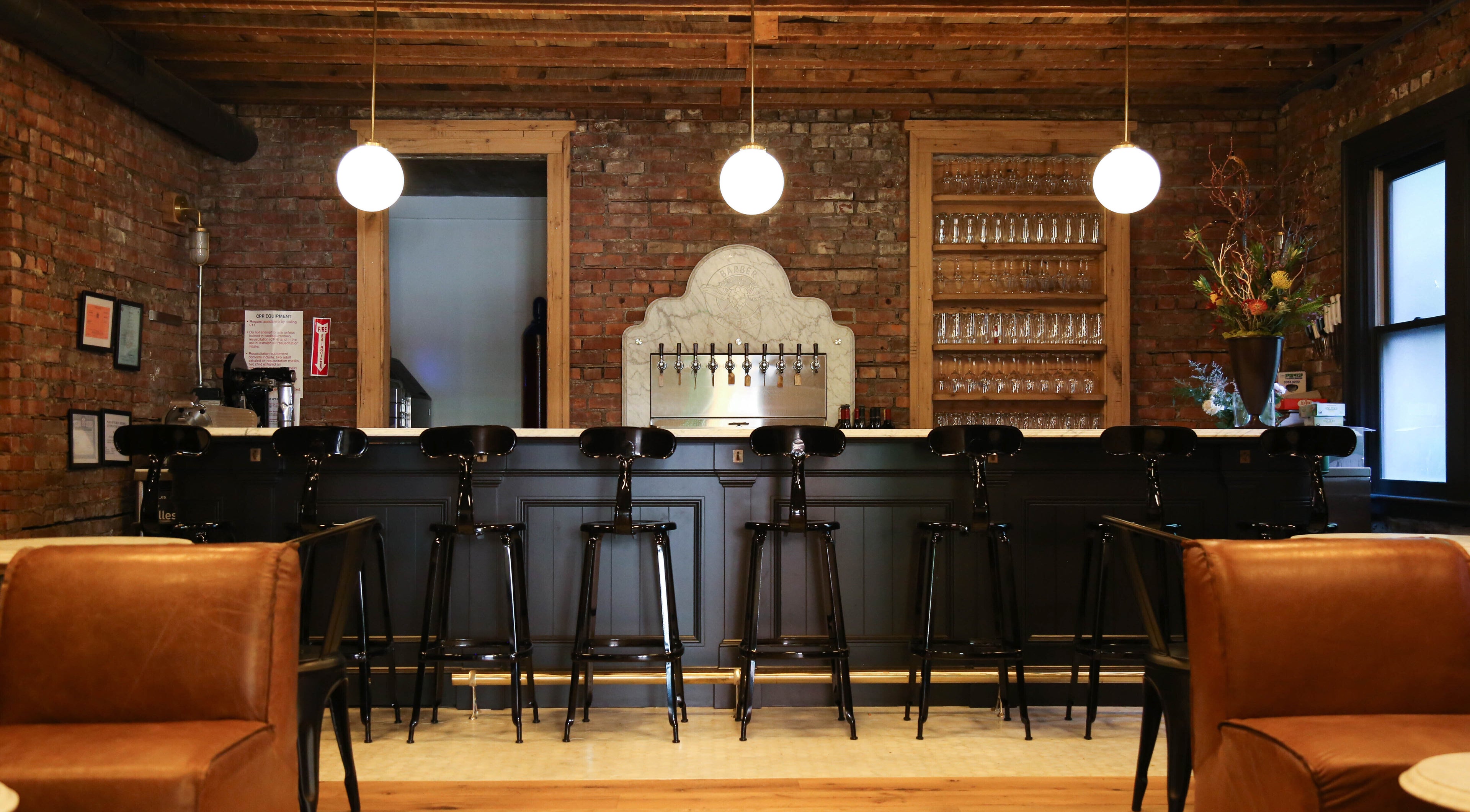 Inviting bar interior at Bar•Ber in Cold Spring, New York, US with a rustic brick backdrop and modern chairs.