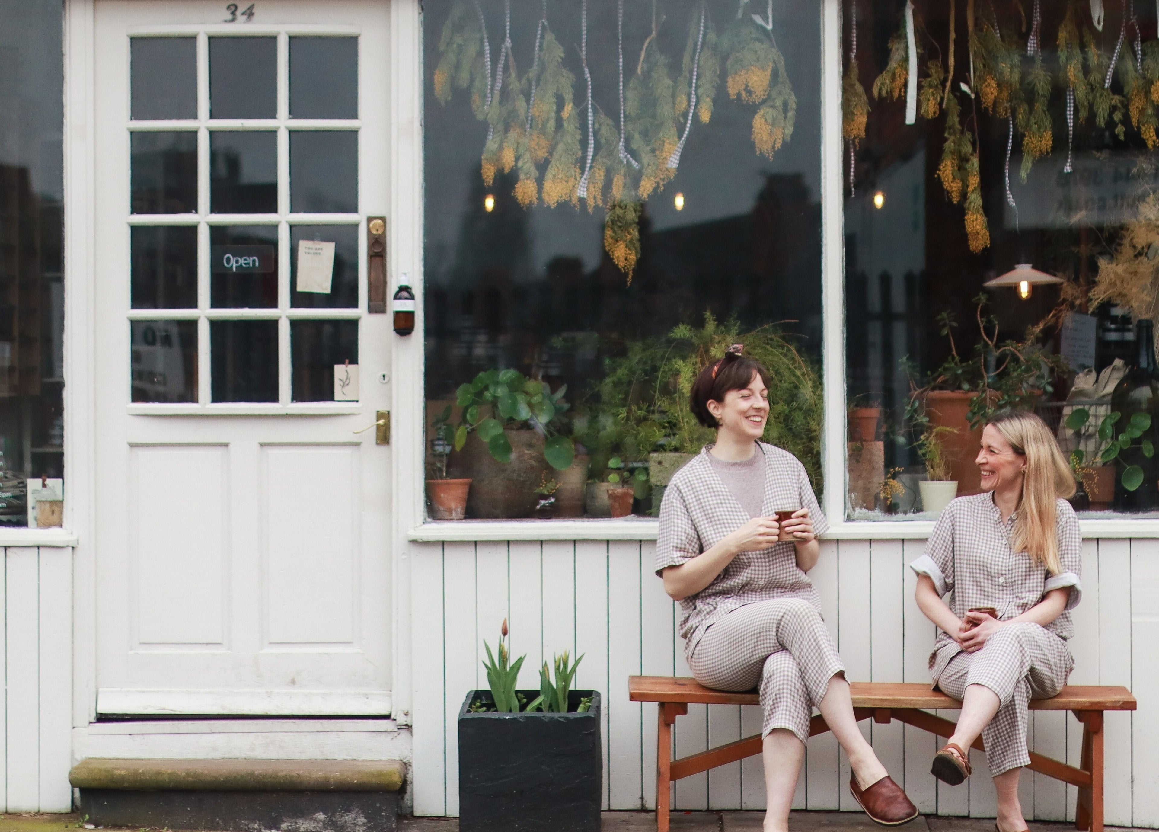 Two women enjoying coffee outside Harvest Skincare, Birmingham, England, GB, surrounded by plants.