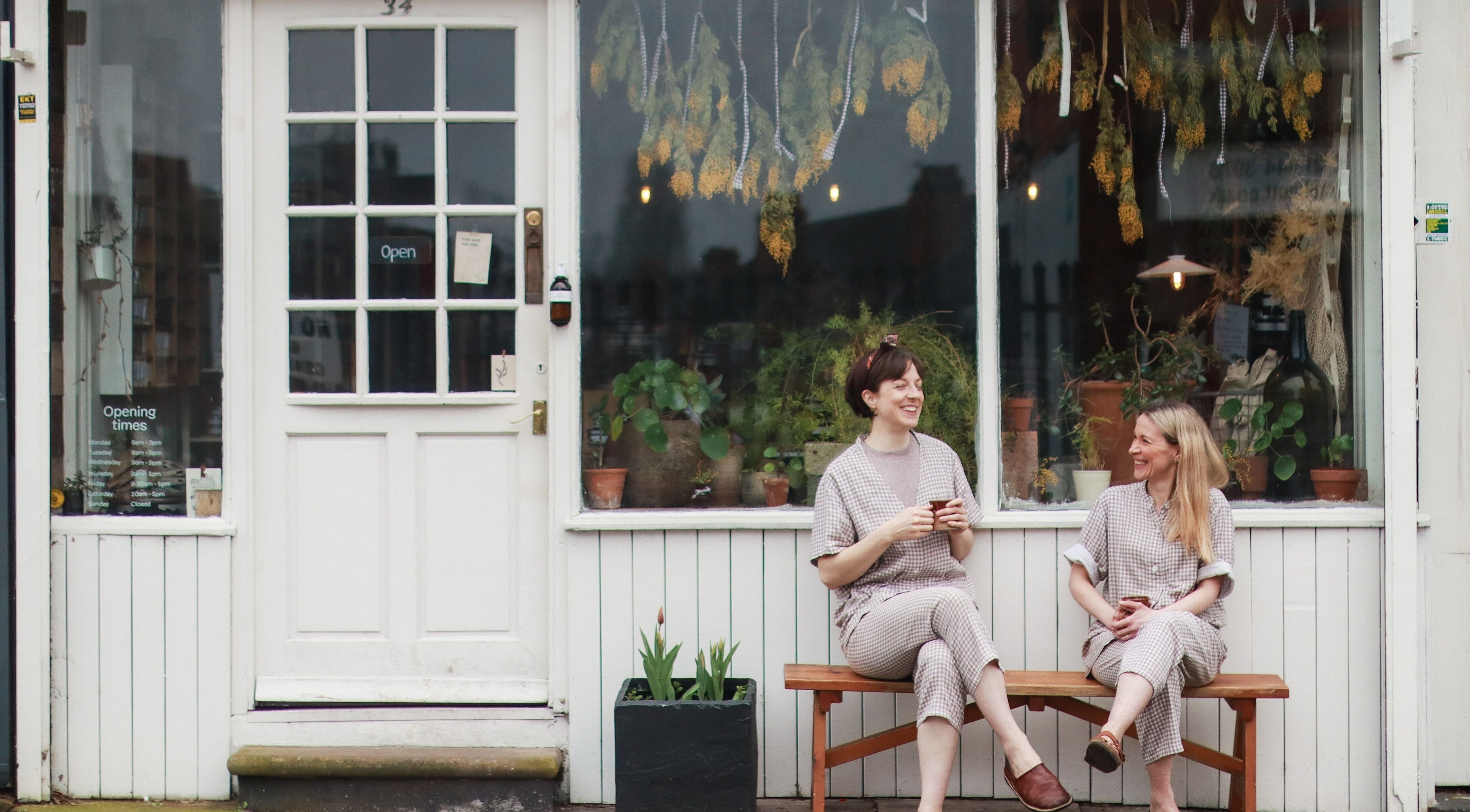 Two women enjoying coffee outside Harvest Skincare, Birmingham, England, GB, surrounded by plants.