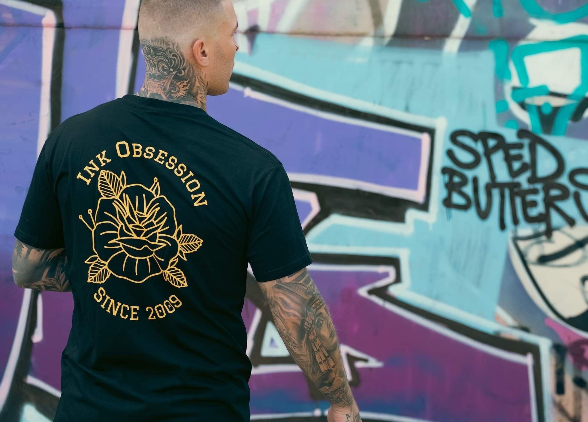 Man in Ink Obsession Tattoo & Piercing shirt by graffiti wall in Nuriootpa, South Australia, AU.
