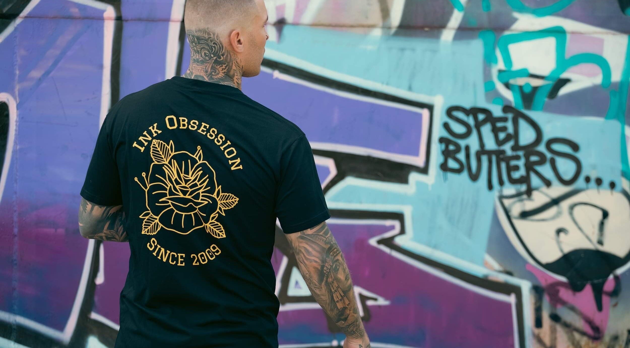 Man in Ink Obsession Tattoo & Piercing shirt by graffiti wall in Nuriootpa, South Australia, AU.
