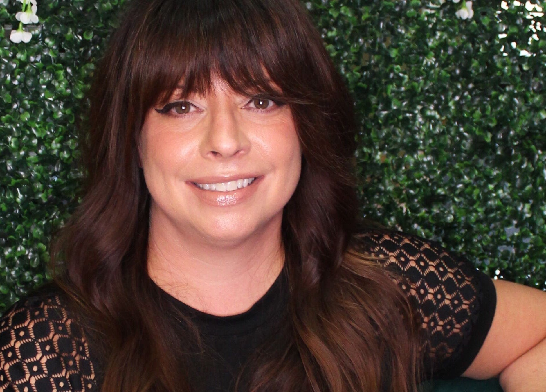 Smiling woman at Fringe Salon, Elkhart, Indiana, US, with lush greenery background.