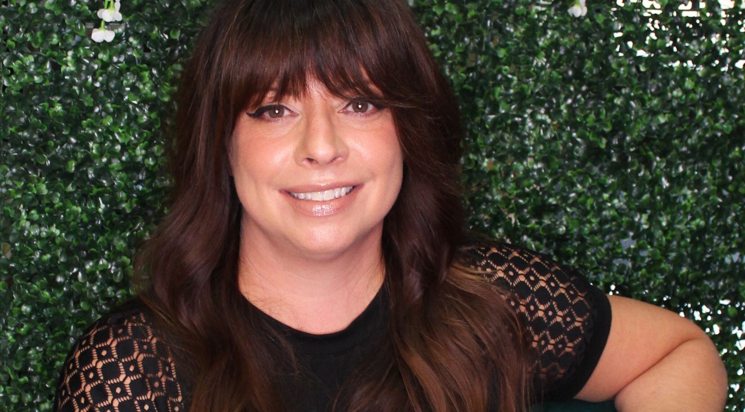 Smiling woman at Fringe Salon, Elkhart, Indiana, US, with lush greenery background.