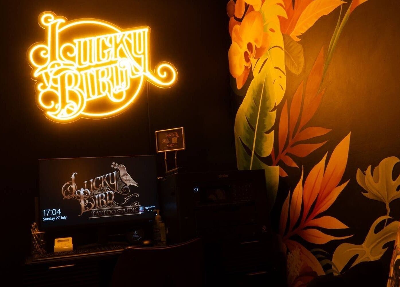 Vibrant neon sign in Lucky Bird Tattoo Studio, Somerton, England, GB with artistic mural and workspace.