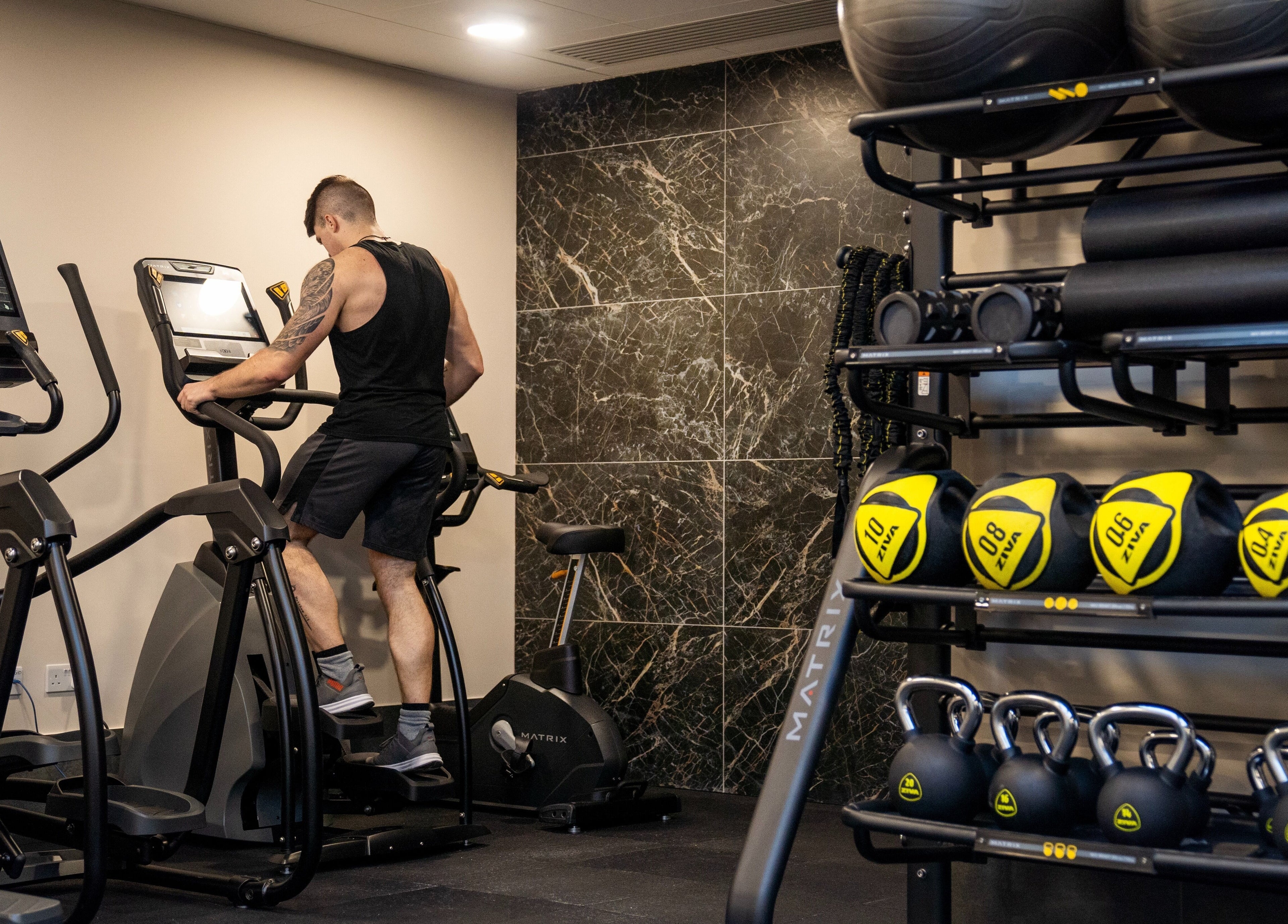 A person exercises on an elliptical at E1 Spa and Wellness, Gibraltar, GI, with kettlebells and gym balls visible.