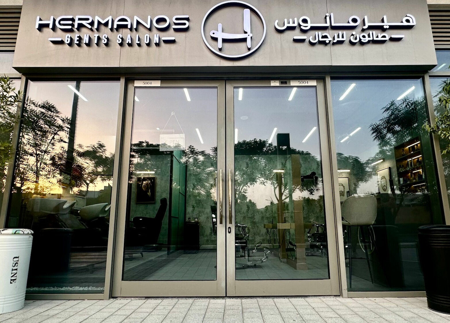Entrance of Hermanos Gents Salon in Dubai, AE with modern glass doors at dusk.