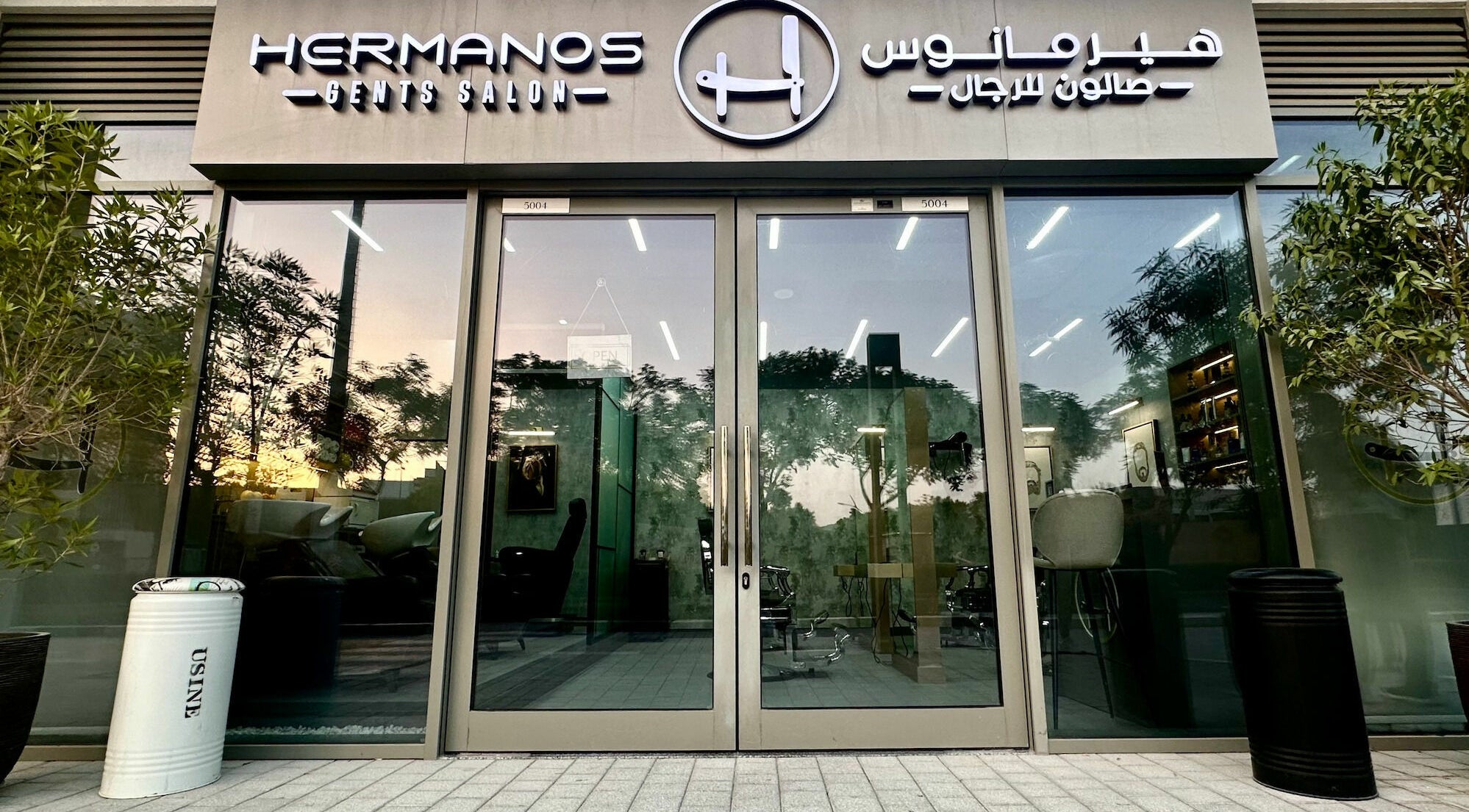 Entrance of Hermanos Gents Salon in Dubai, AE with modern glass doors at dusk.