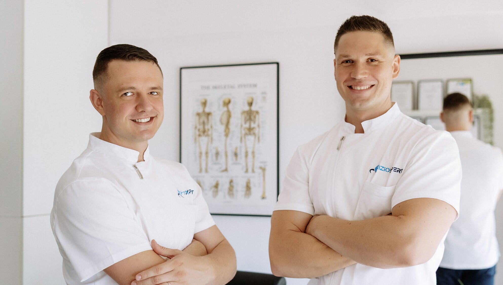 Dva terapeuta iz Fizio Expert Zagreba, Grad Zagreb, HR, u bijelim uniformama pred anatomskim posterom.