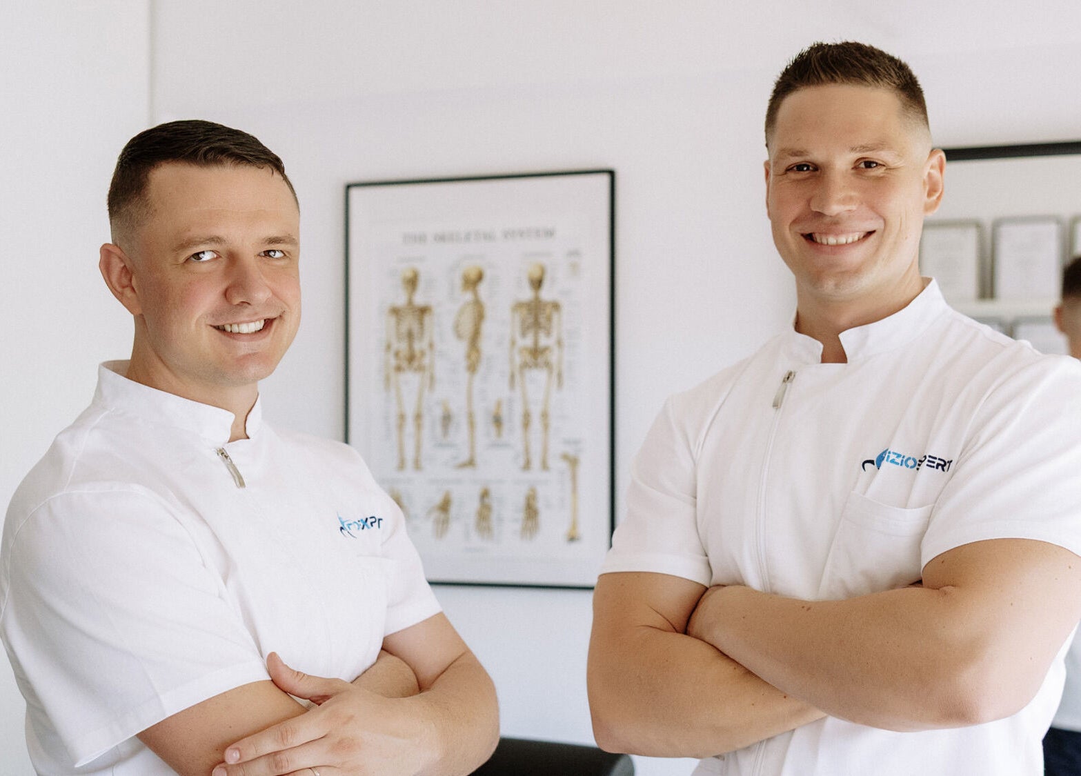 Dva terapeuta iz Fizio Expert Zagreba, Grad Zagreb, HR, u bijelim uniformama pred anatomskim posterom.