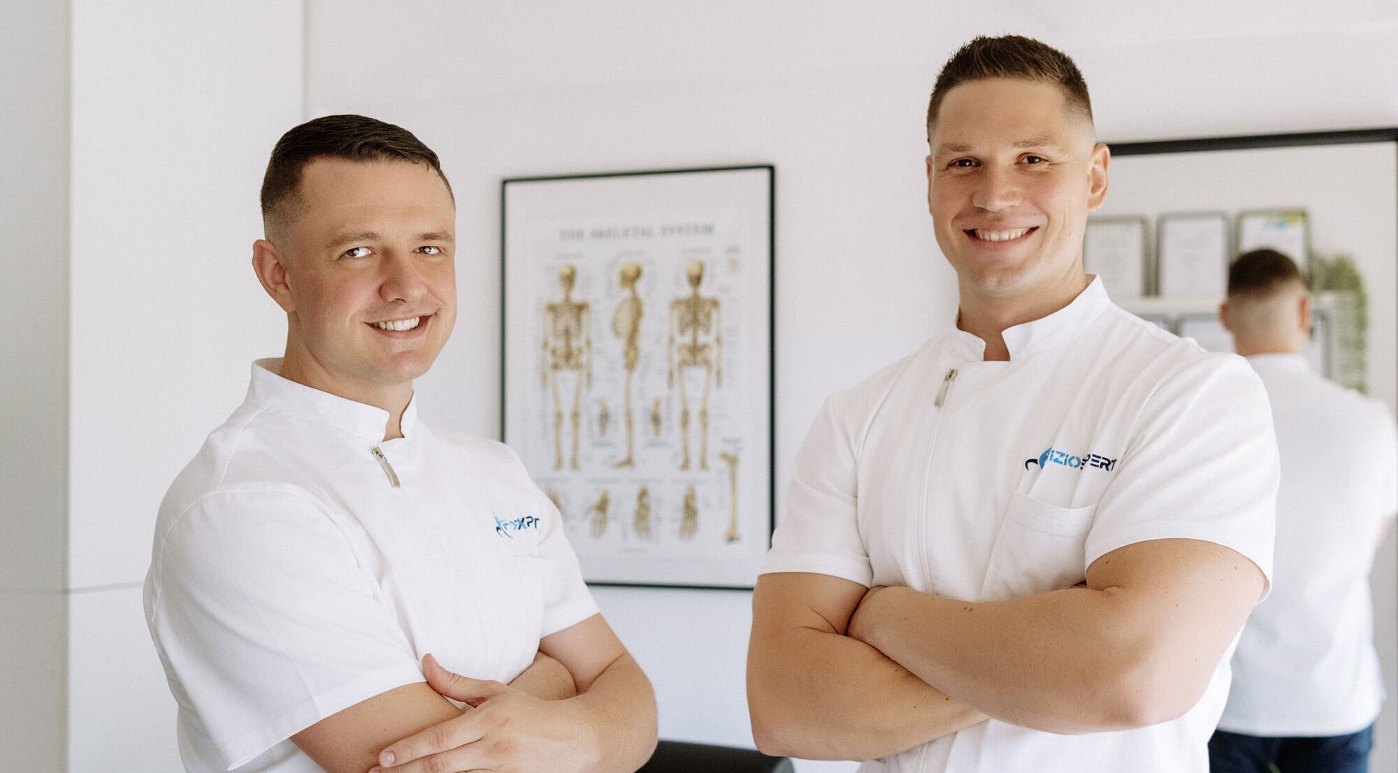 Dva terapeuta iz Fizio Expert Zagreba, Grad Zagreb, HR, u bijelim uniformama pred anatomskim posterom.