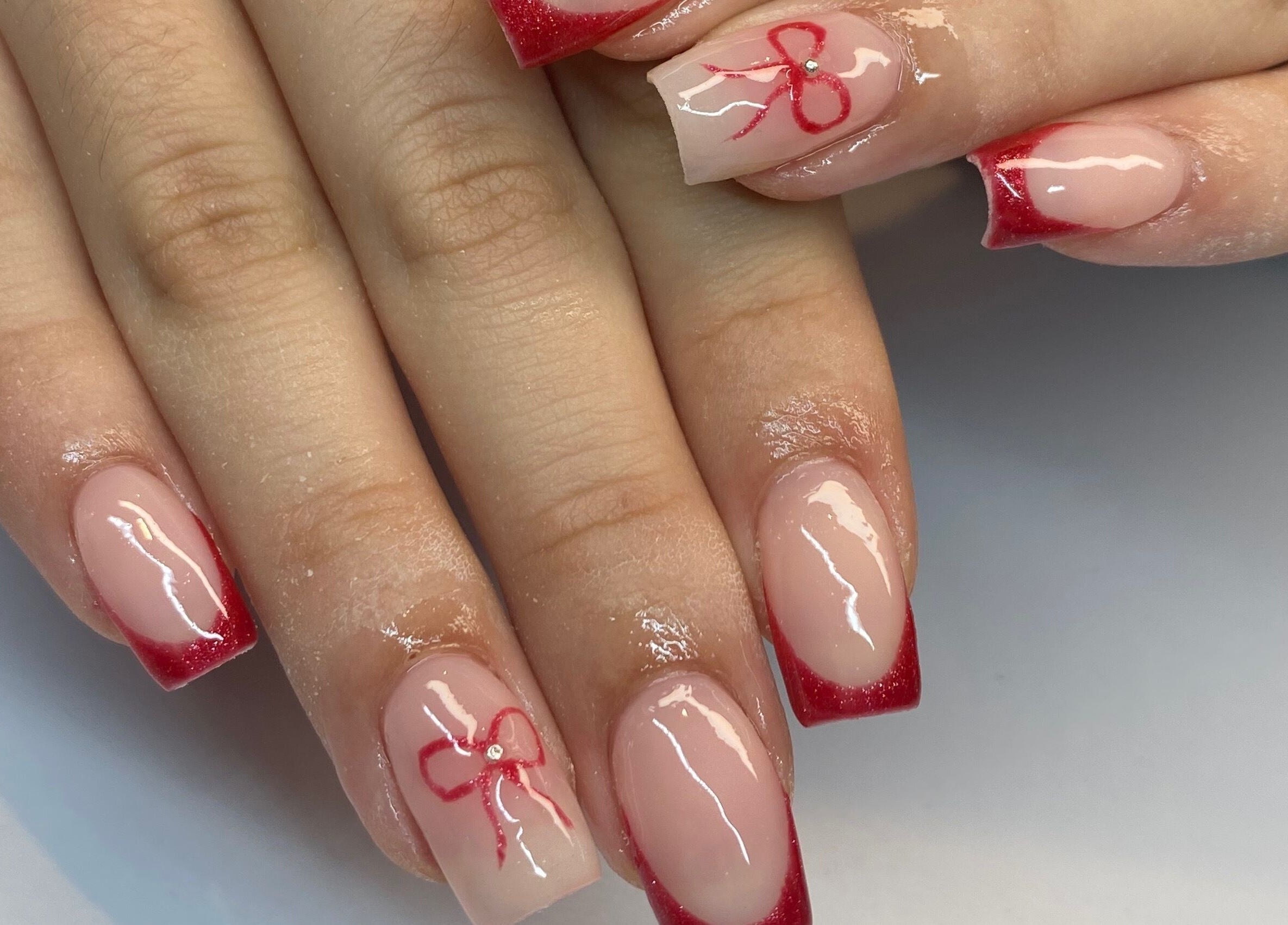 Stylish red-tipped nails with bow design at Polished By Jules in Caledon, Ontario, CA. Trendy and elegant manicure.