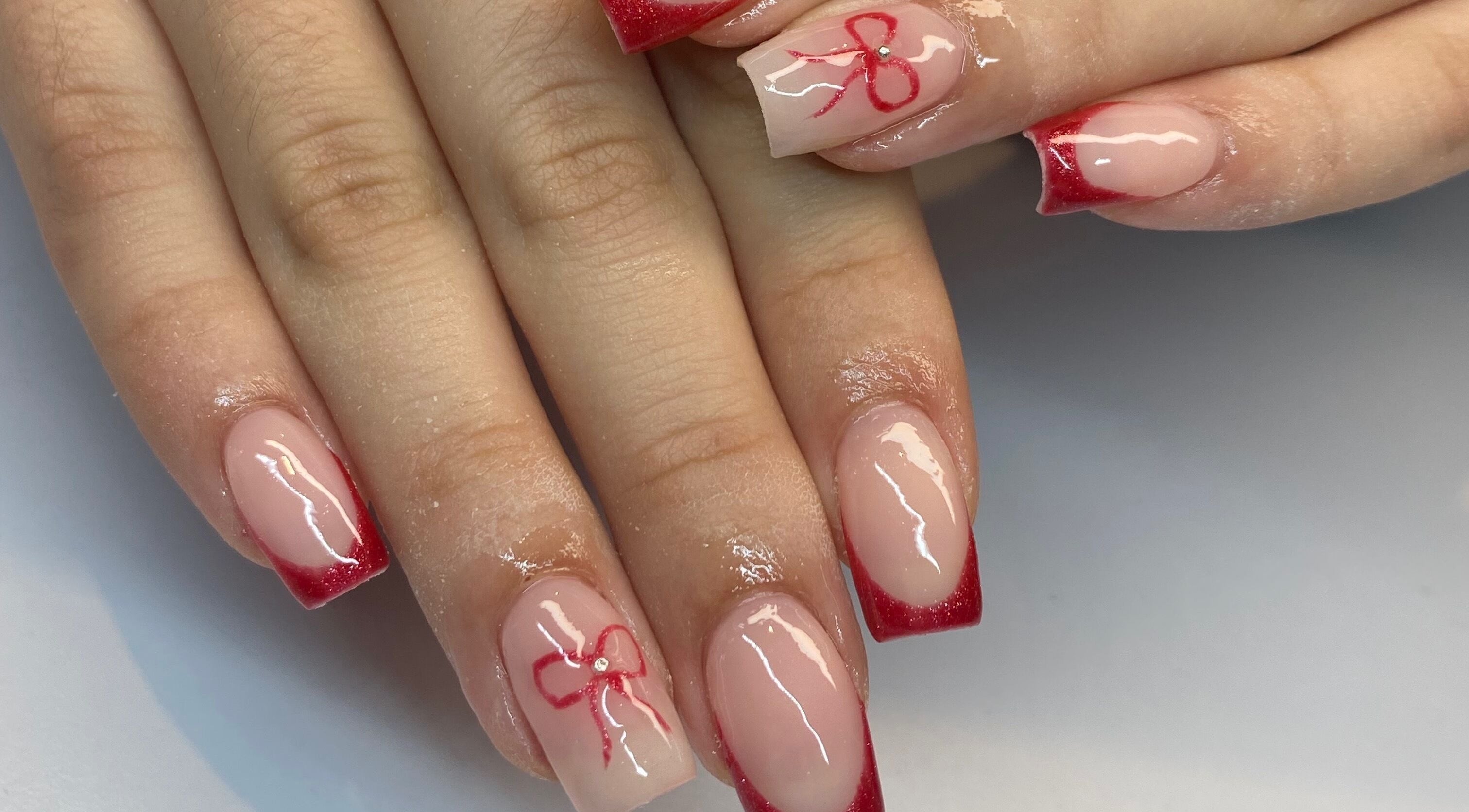 Stylish red-tipped nails with bow design at Polished By Jules in Caledon, Ontario, CA. Trendy and elegant manicure.