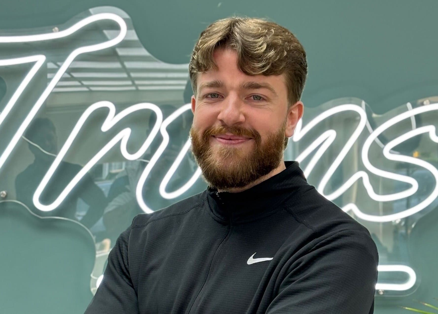 Smiling barber at Bez Barber, Birmingham, England, GB, standing confidently in front of neon sign.