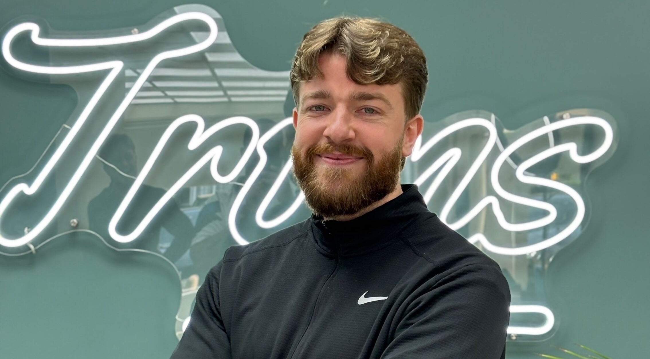 Smiling barber at Bez Barber, Birmingham, England, GB, standing confidently in front of neon sign.
