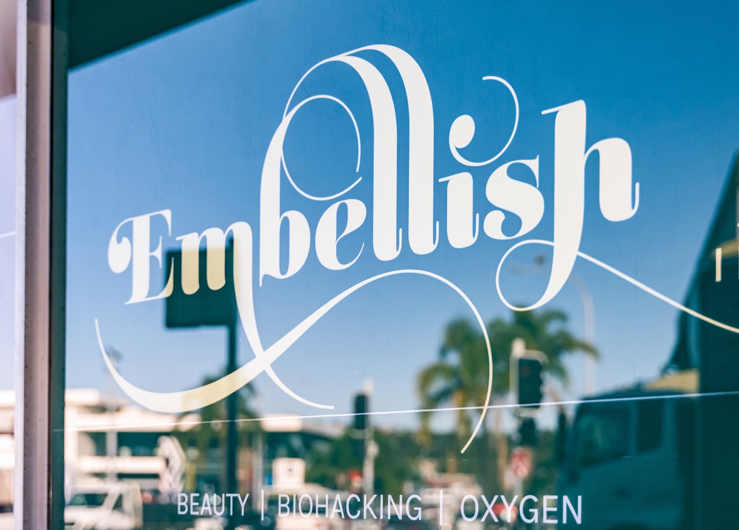 Window signage of Embellish Inc at Brookvale, NSW, AU showcasing stylish logo with blue sky reflection.
