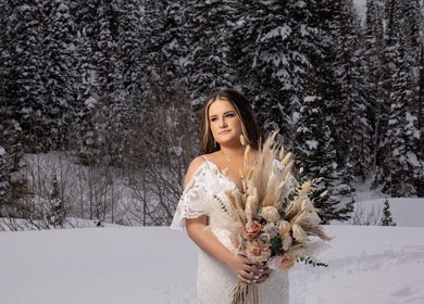 Bride in snow, holding bouquet at Meg You Glow in Heber City, Utah, US, surrounded by snow-covered trees.