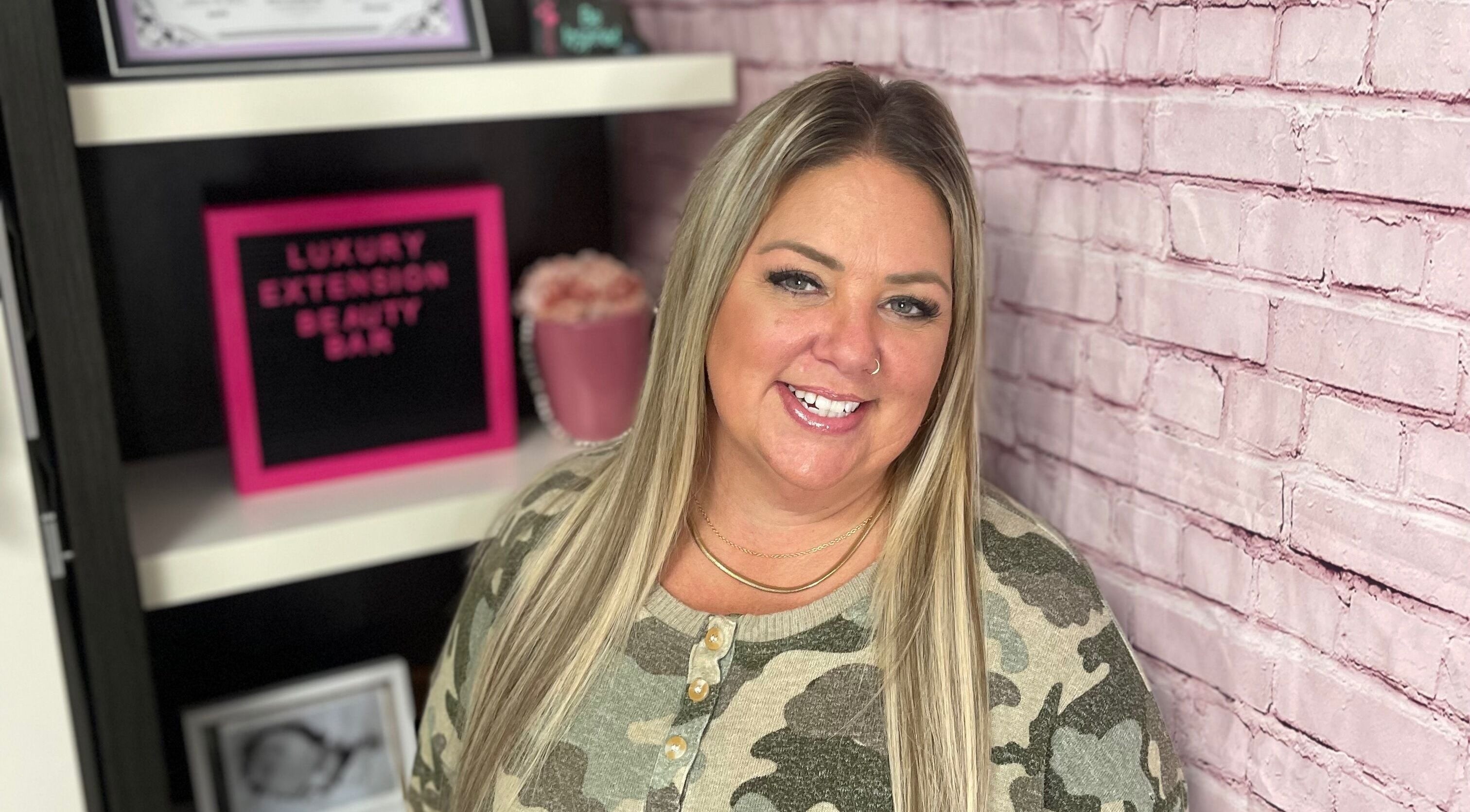 Professional at Orillia & Area Luxury Extension Beauty Bar, Orillia, Ontario, CA, smiling by pink brick wall.